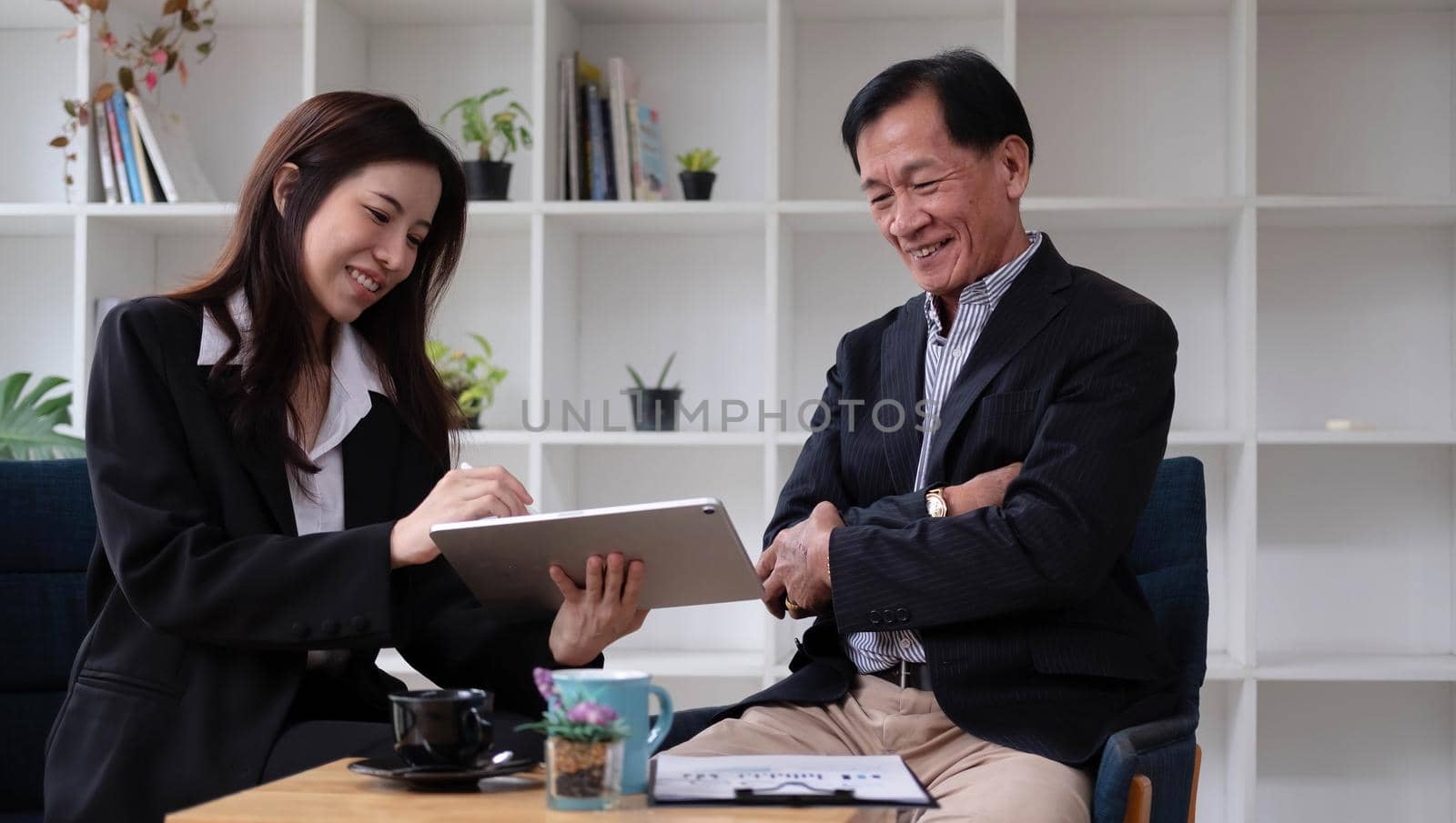 Happy woman and middle-aged man sit at office desk work on laptop together laughing on funny joke, smiling diverse colleagues have fun talking cooperating at workplace by wichayada