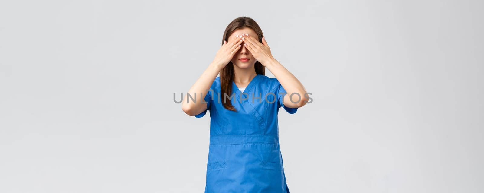 Healthcare workers, prevent virus, insurance and medicine concept. Young woman in blue scrubs, doctor or nurse close eyes with hands, blindfolded medicine, standing grey background.