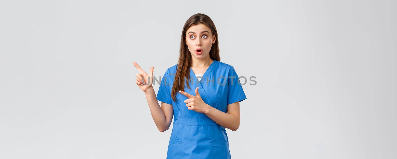 Healthcare workers, prevent virus, insurance and medicine concept. Surprised female nurse or doctor in blue scrubs, looking and pointing fingers upper left corner, gasping amazed.