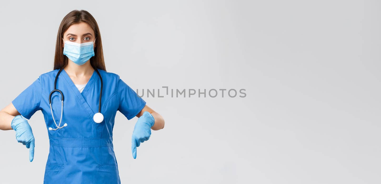 Covid-19, preventing virus, health, healthcare workers and quarantine concept. Determined female nurse or doctor in blue scrubs, medical mask and gloves, pointing fingers down, showing info banner.