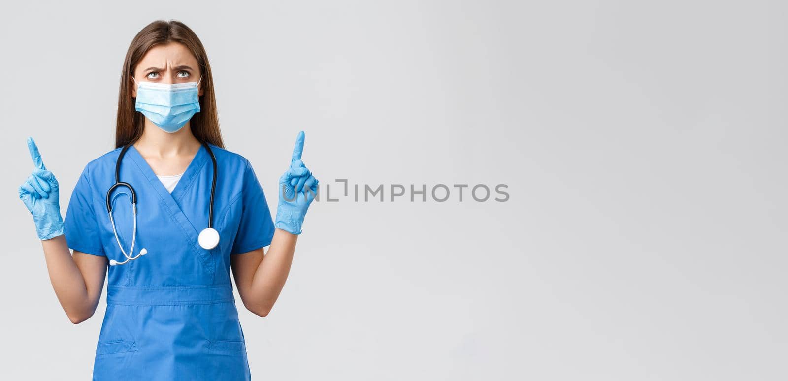 Covid-19, preventing virus, health, healthcare workers and quarantine concept. Concerned and disappointed female nurse or doctor in blue scrubs, medical mask, frowning looking up displeased.