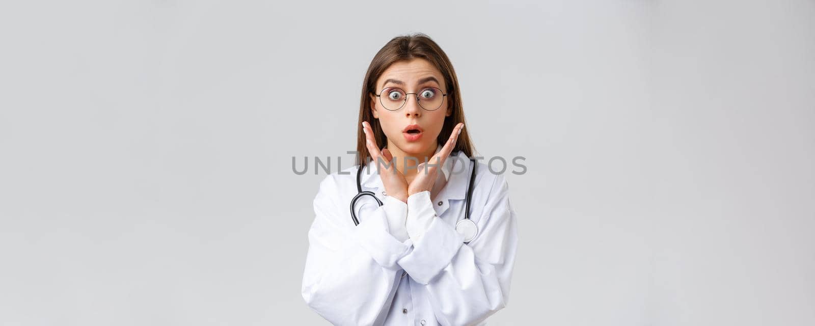 Healthcare workers, medicine, insurance and covid-19 pandemic concept. Surprised and impressed female doctor in glasses, white scrubs, gasping and staring astonished camera, grey background.