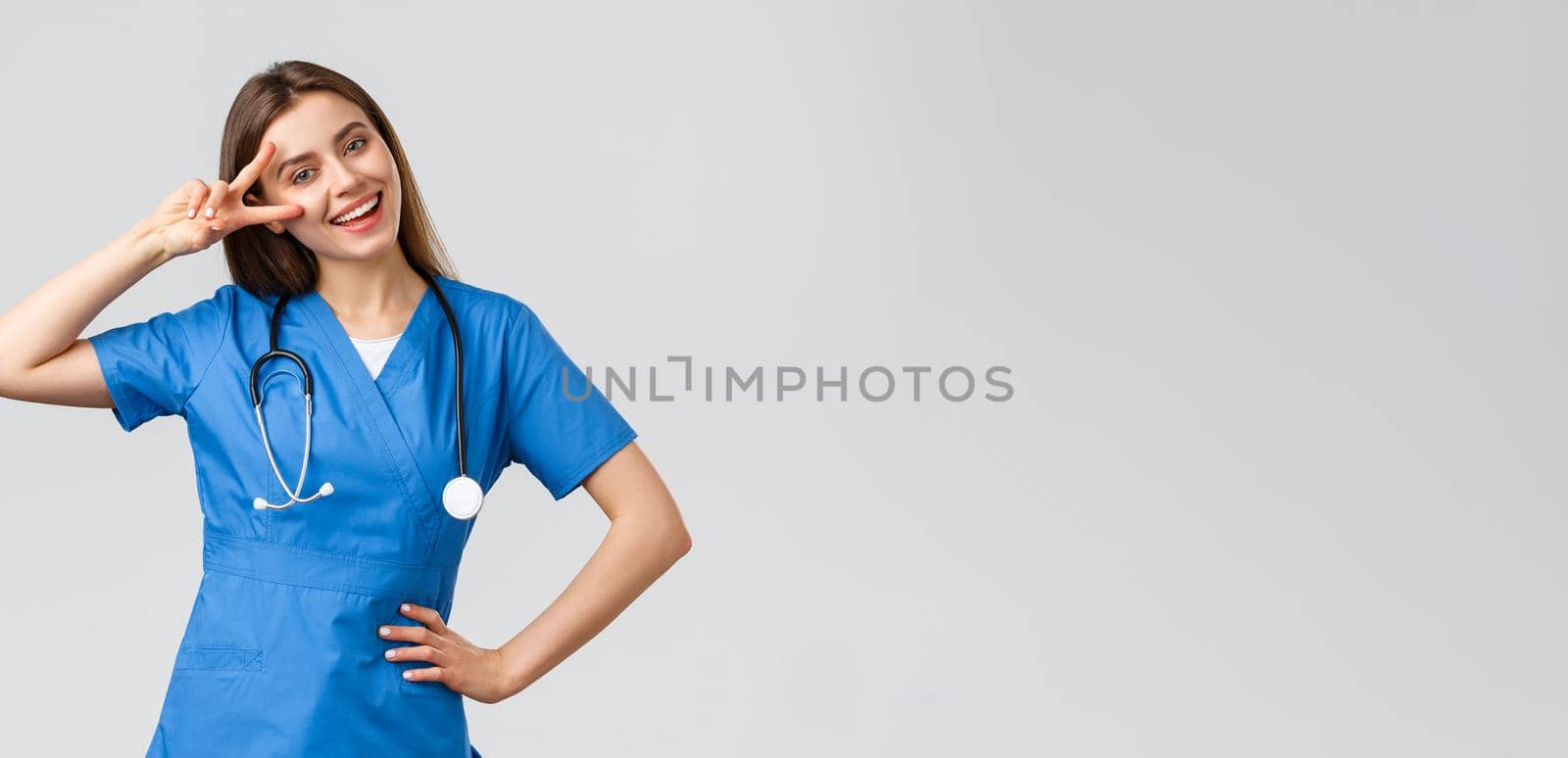 Medical workers, healthcare, covid-19 and vaccination concept. Cheerful attractive female nurse or doctor in blue scrubs, stethoscope, show peace sign, stay optimistic, standing grey background.