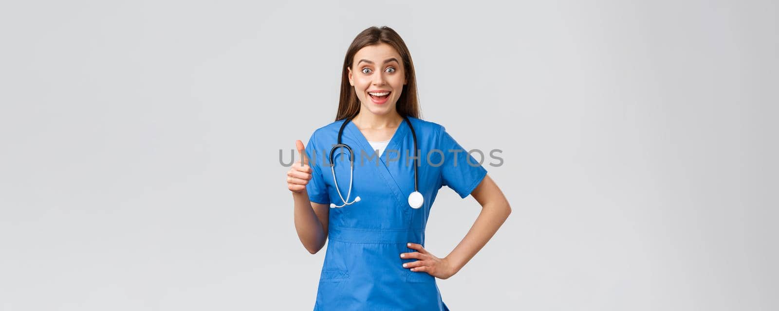 Medical workers, healthcare, covid-19 and vaccination concept. Surprised excited female nurse or doctor in blue scrubs with stethoscope, show thumbs-up in approval, like idea, smiling upbeat.