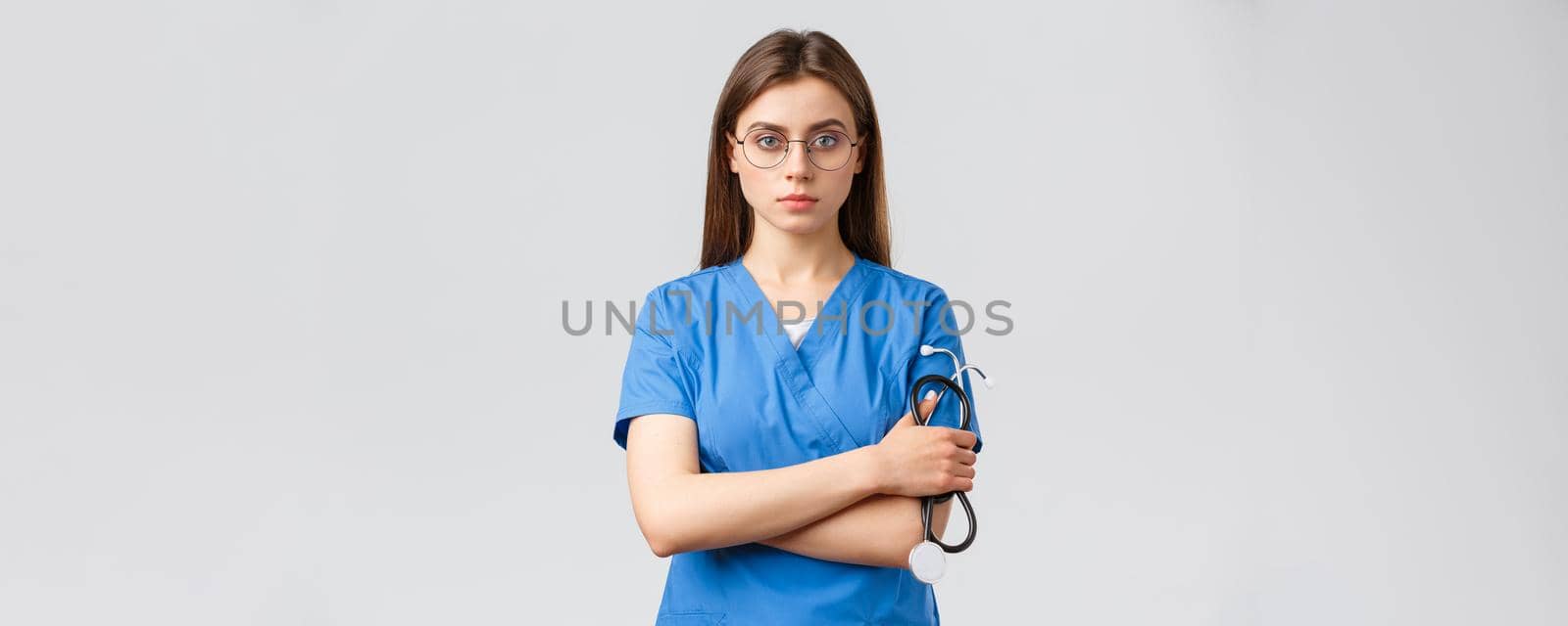 Healthcare workers, medicine, insurance and covid-19 pandemic concept. Serious and determined, professional female nurse, doctor in blue scrubs, holding stethoscope, wear glasses, look confident.