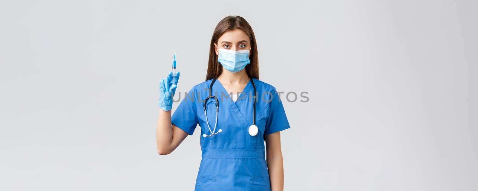 Covid-19, preventing virus, health, healthcare workers and quarantine concept. Serious female nurse in blue scrubs, doctor wearing medical mask holding syringe with coronavirus vaccine.
