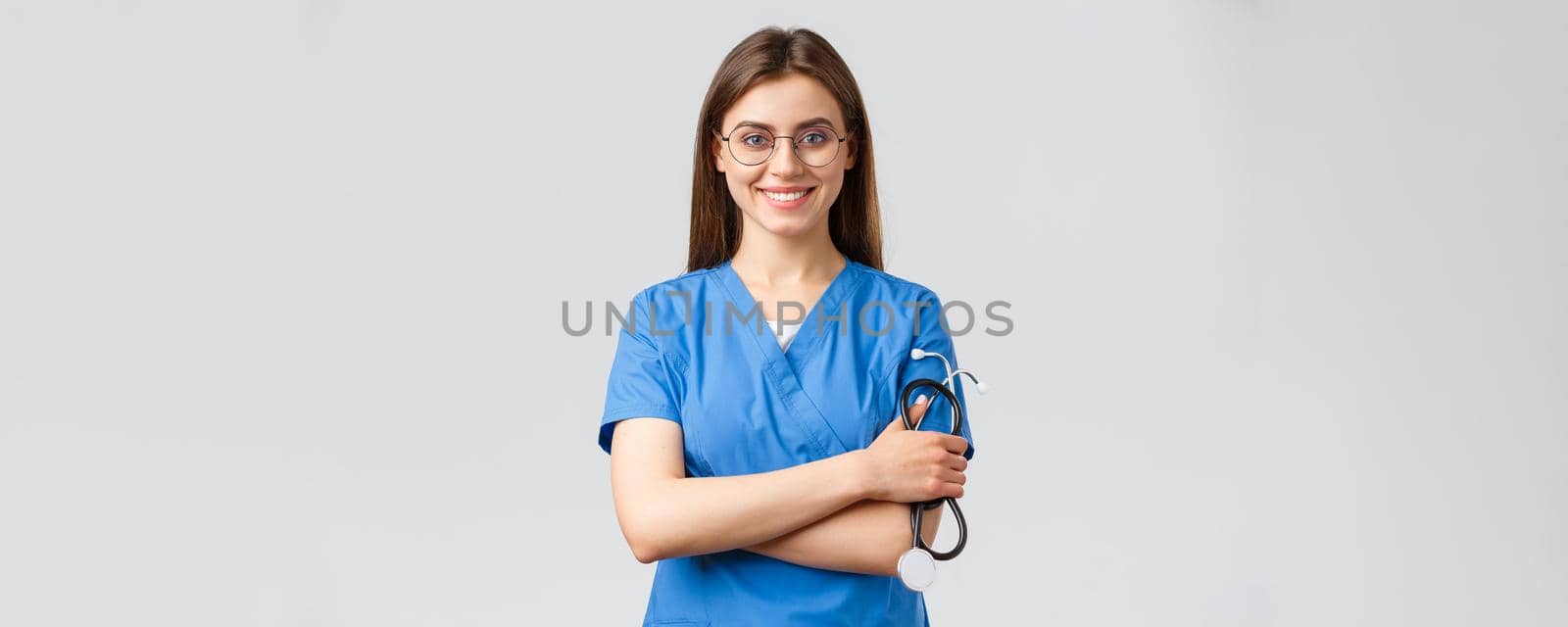 Healthcare workers, medicine, insurance and covid-19 pandemic concept. Confident and enthusiastic young female nurse, doctor in blue scrubs and glasses, hold stethoscope, smiling camera pleased.