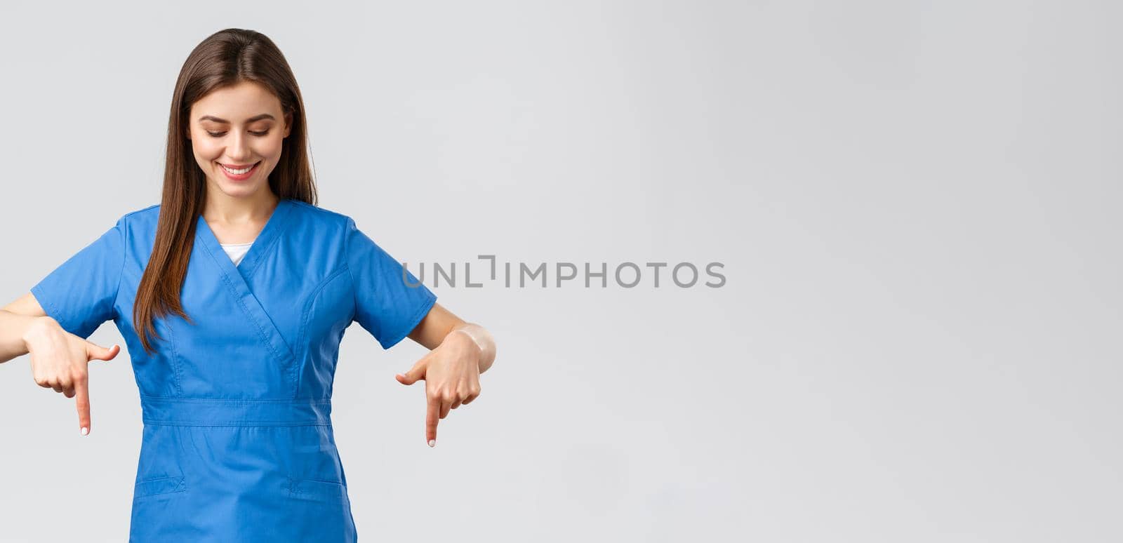 Healthcare workers, prevent virus, covid-19 test screening, medicine concept. Happy and pleased attractive female nurse or doctor, physician in blue scrubs, pointing looking down at advertisement by Benzoix