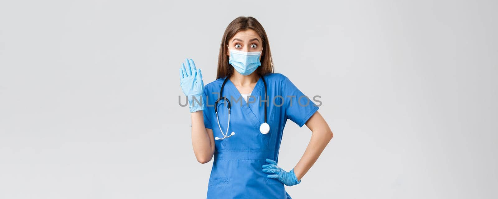 Covid-19, preventing virus, health, healthcare workers and quarantine concept. Surprised cute female nurse in blue scrubs saying hello, see someone in clinic, wave hi, wear medical mask and gloves by Benzoix