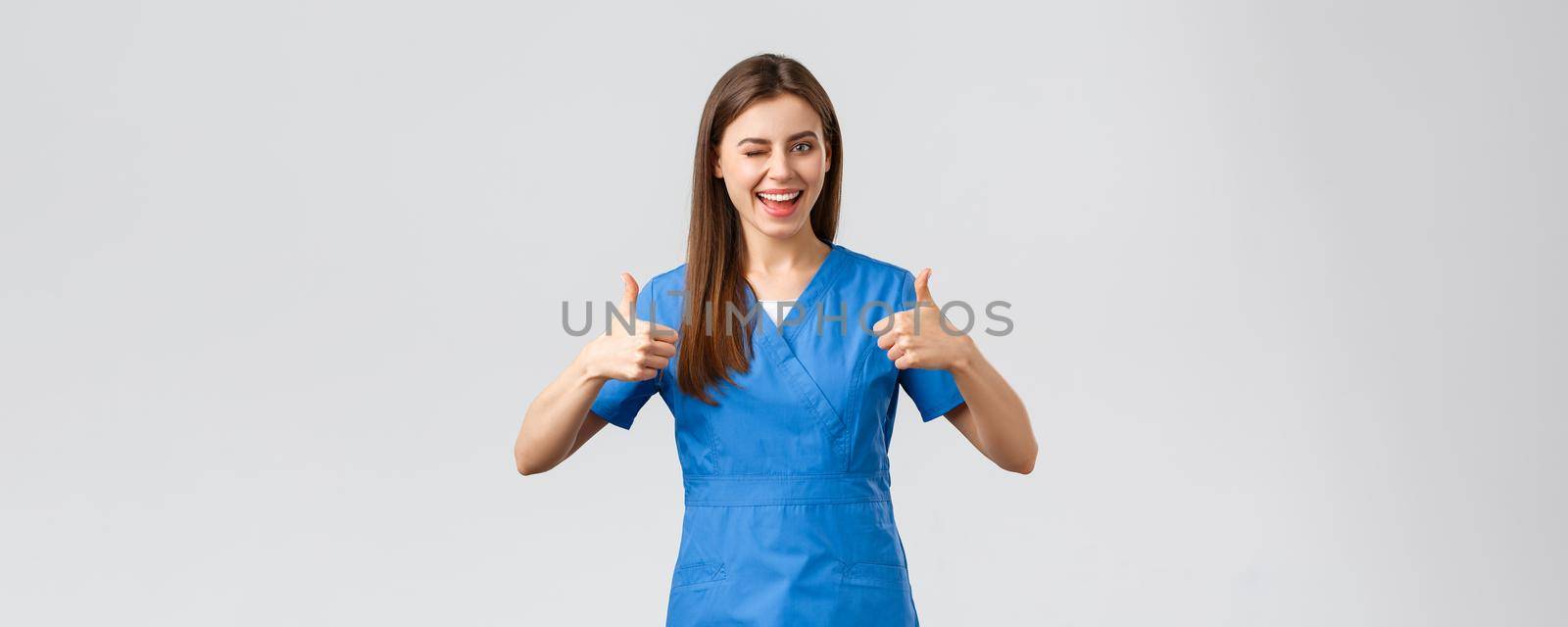 Healthcare workers, prevent virus, insurance and medicine concept. Optimistic good-looking female nurse or doctor in scrubs, wink encourage visit clinic, show thumbs-up in approval.