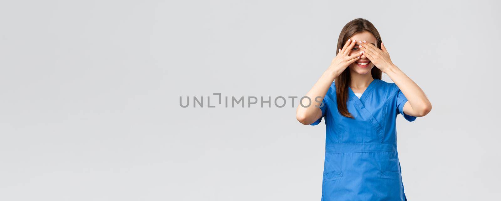 Healthcare workers, prevent virus, insurance and medicine concept. Cute female nurse or doctor in clinic wearing scrubs, cover eyes and peeking through fingers with intrigued smile.