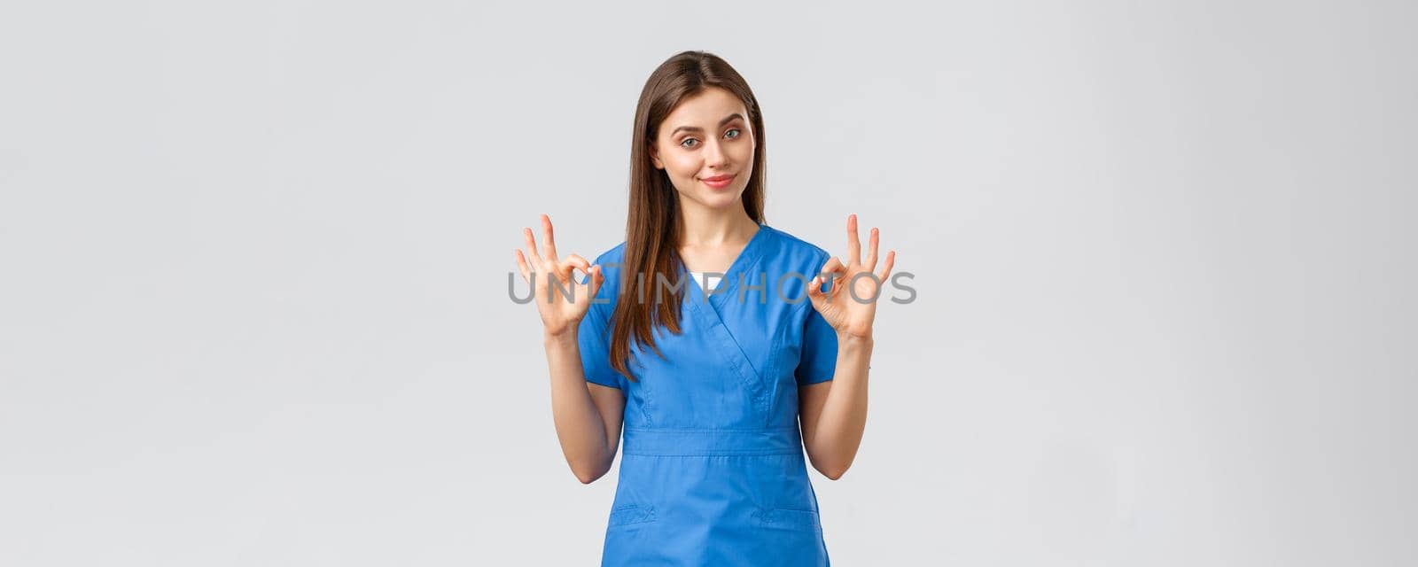 Healthcare workers, prevent virus and medicine concept. Attractive confident female nurse, doctor in blue scrubs, smirk show okay sign, guarantee quality of service in clinic, recommend medicine by Benzoix