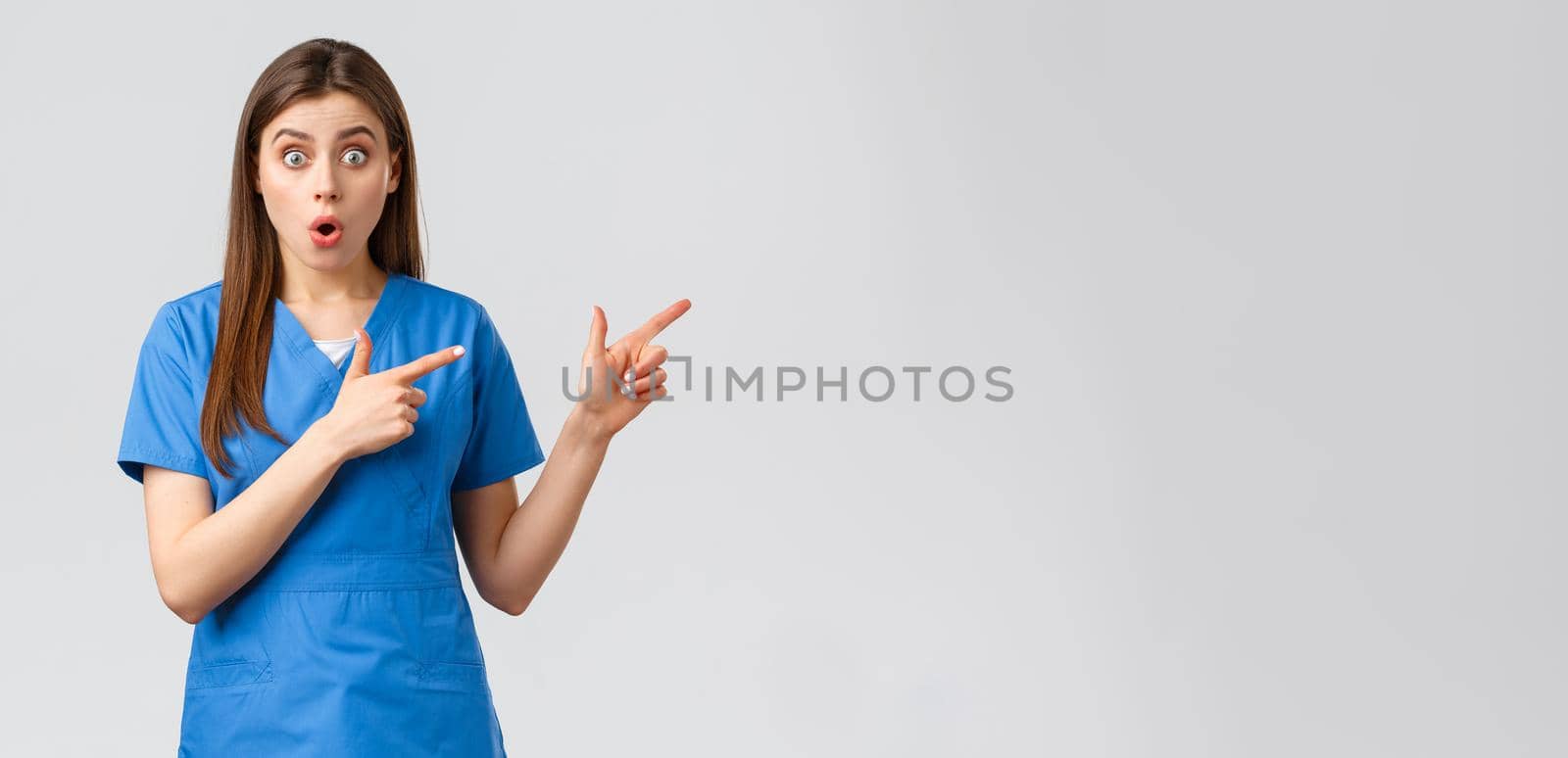 Healthcare workers, prevent virus, insurance and medicine concept. Surprised nurse or doctor in blue scrubs listen to interesting news, pointing fingers right, gasping shocked at camera.