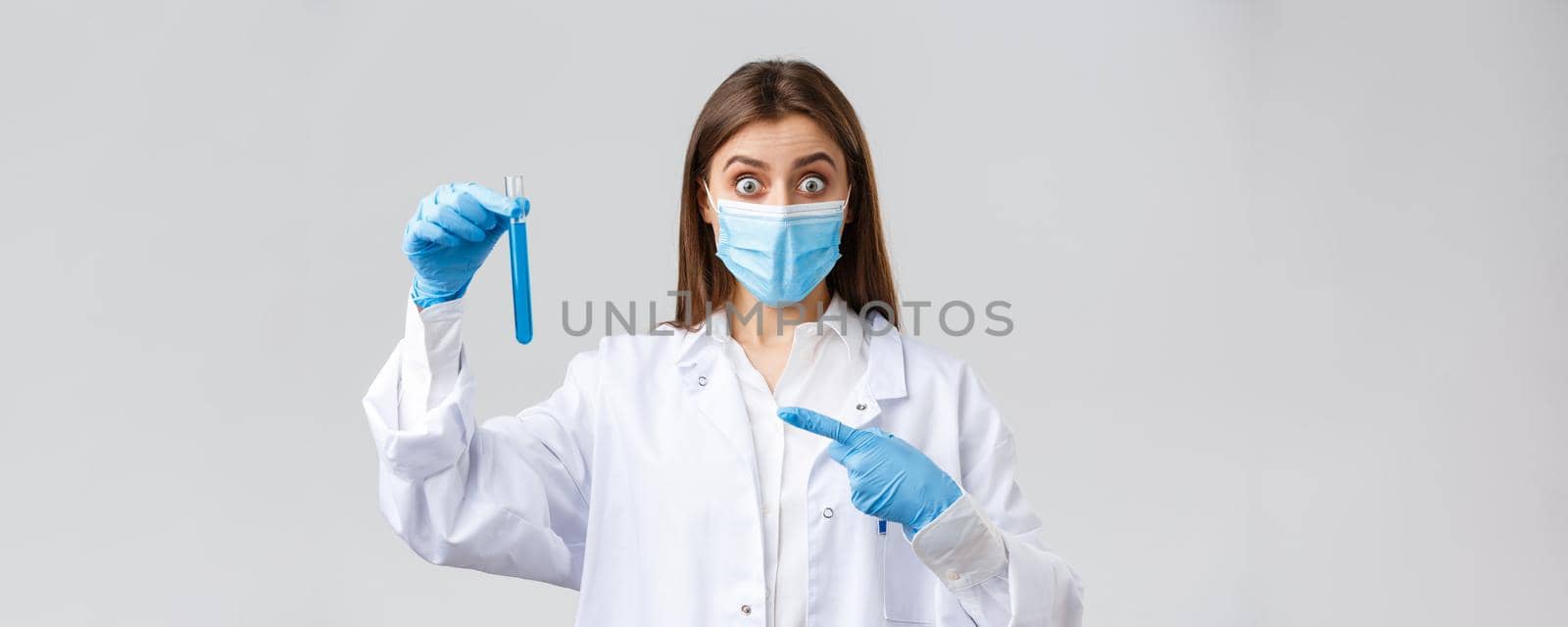 Covid-19, preventing virus, healthcare workers concept. Excited and surprised doctor in medical mask, gloves and scrubs pointing finger at test-tube coronavirus vaccine, impressive sample.