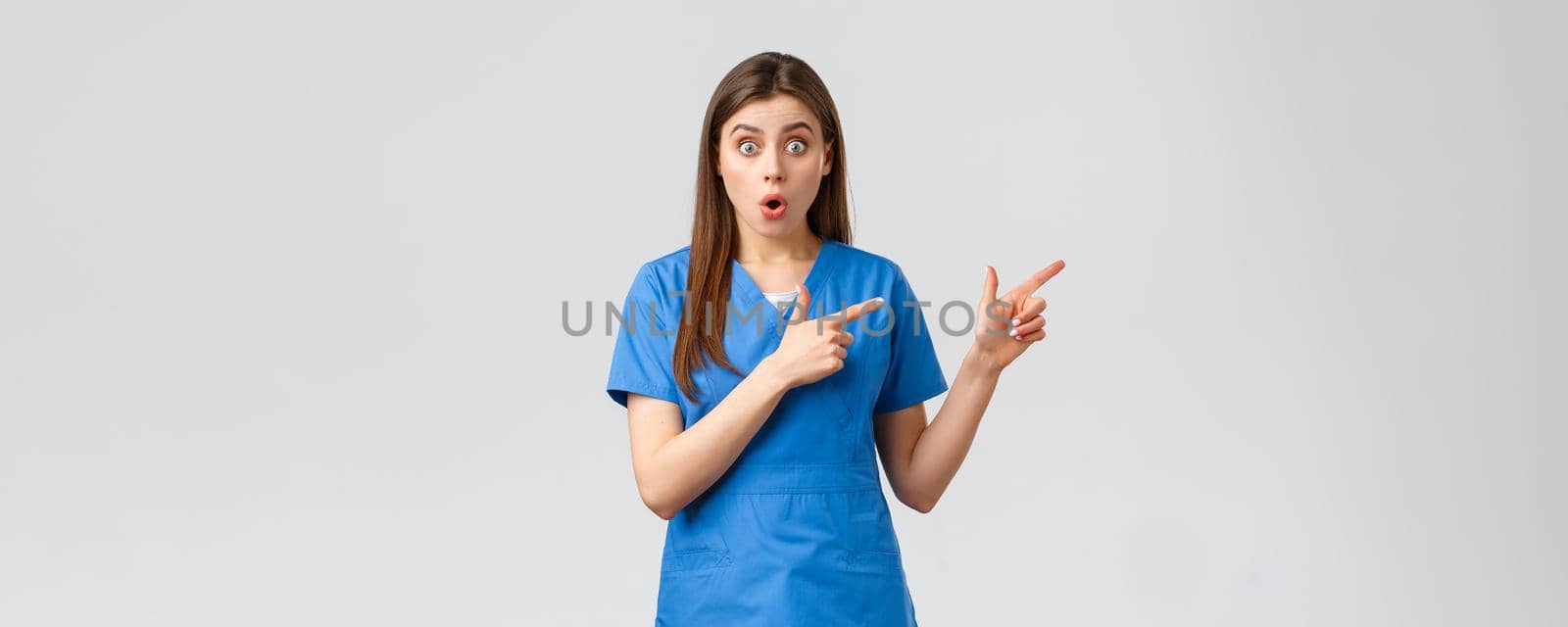 Healthcare workers, prevent virus, insurance and medicine concept. Surprised nurse or doctor in blue scrubs listen to interesting news, pointing fingers right, gasping shocked at camera.