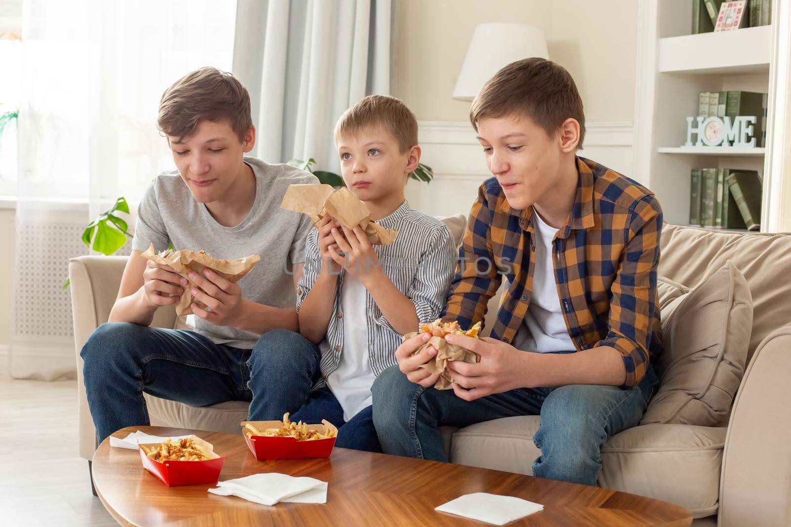 Three happy teenagers, in casual clothes, are sitting on the sofa in the living room, eating fast food, a hamburger and french fries in red boxes.