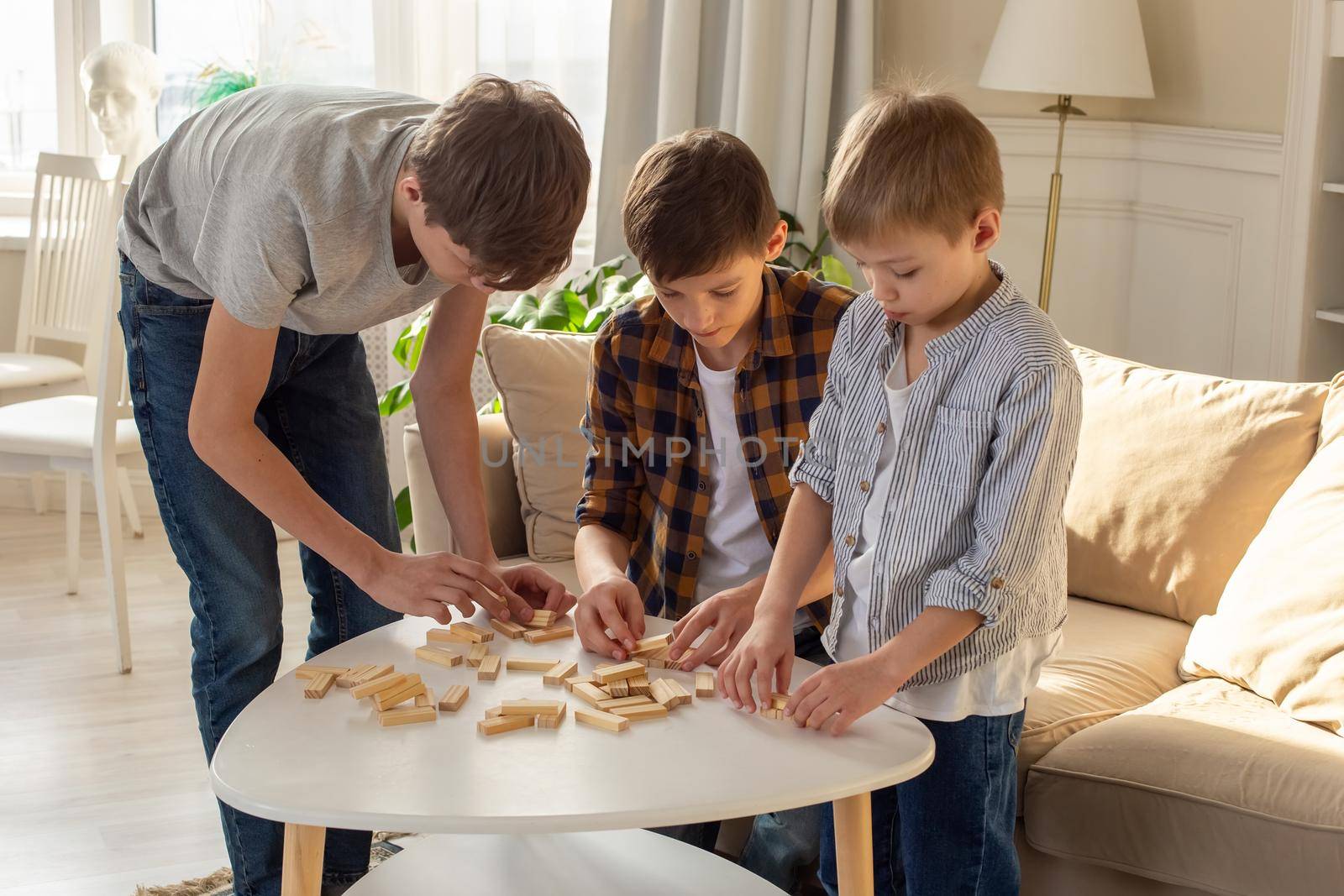Three boys, in a sunny room, are enthusiastically playing a board game made of wooden rectangular blocks, pulling pieces out of the tower. Close-up