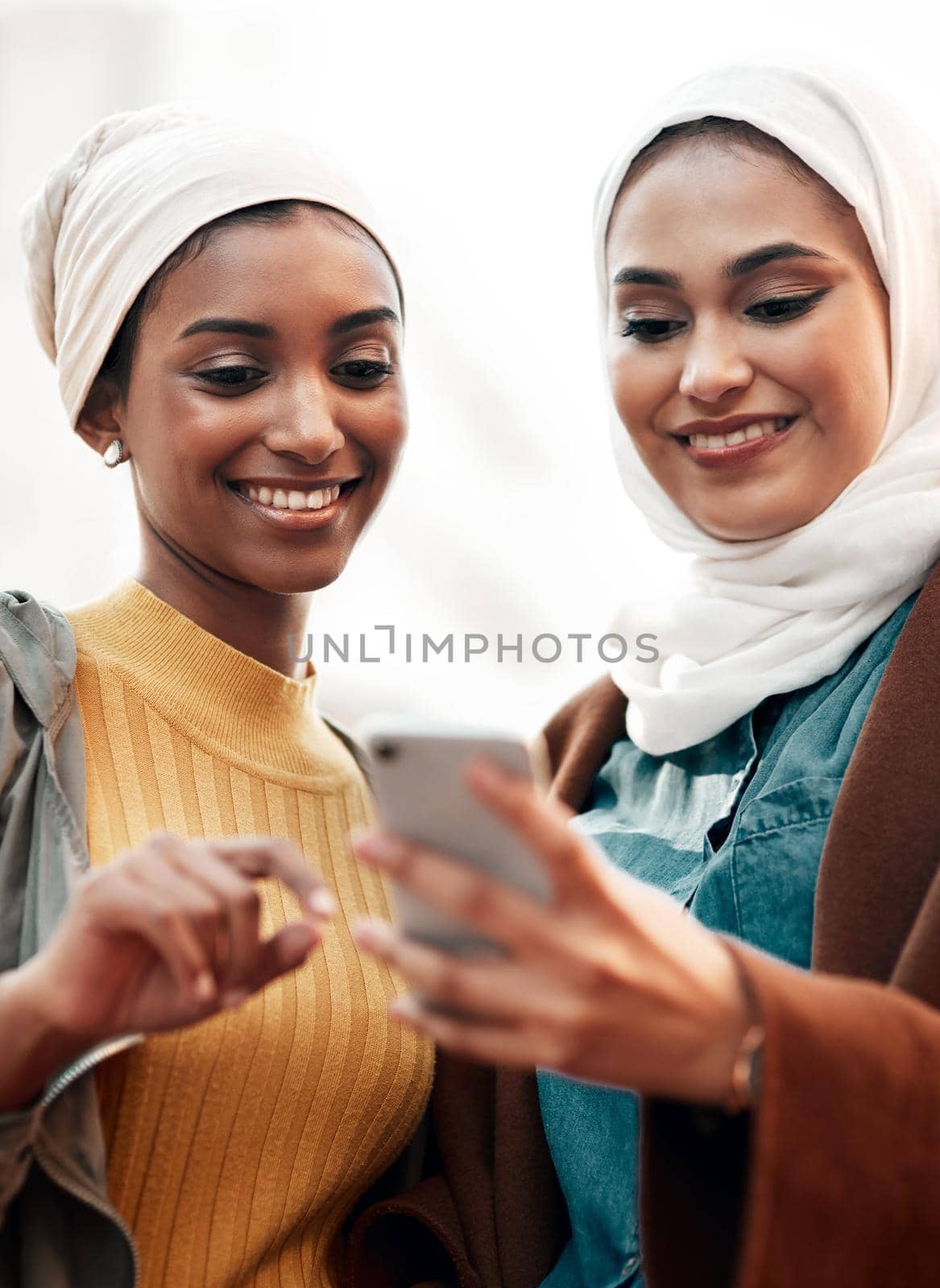 This looks like a nice venue. Cropped shot of two attractive young women wearing headscarves and standing together while using a cellphone in the city. by YuriArcurs