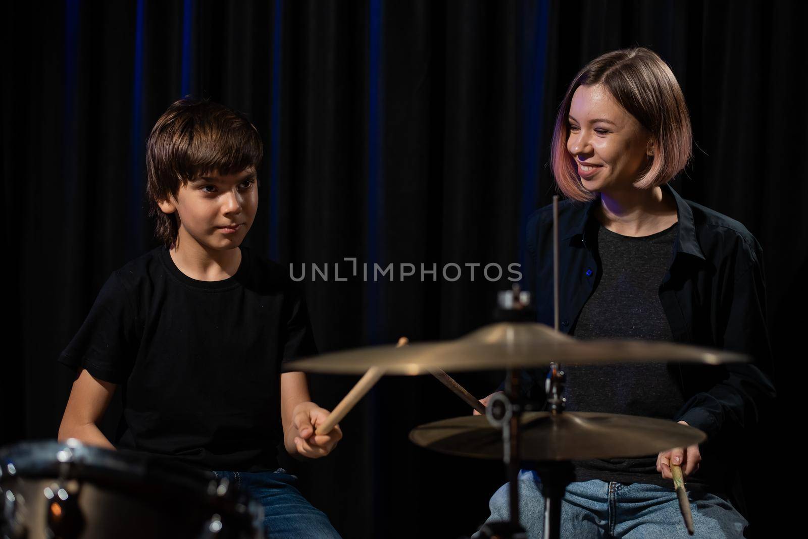 Young caucasian woman teaches a boy to play the drums in the studio on a black background. Music school student by mrwed54