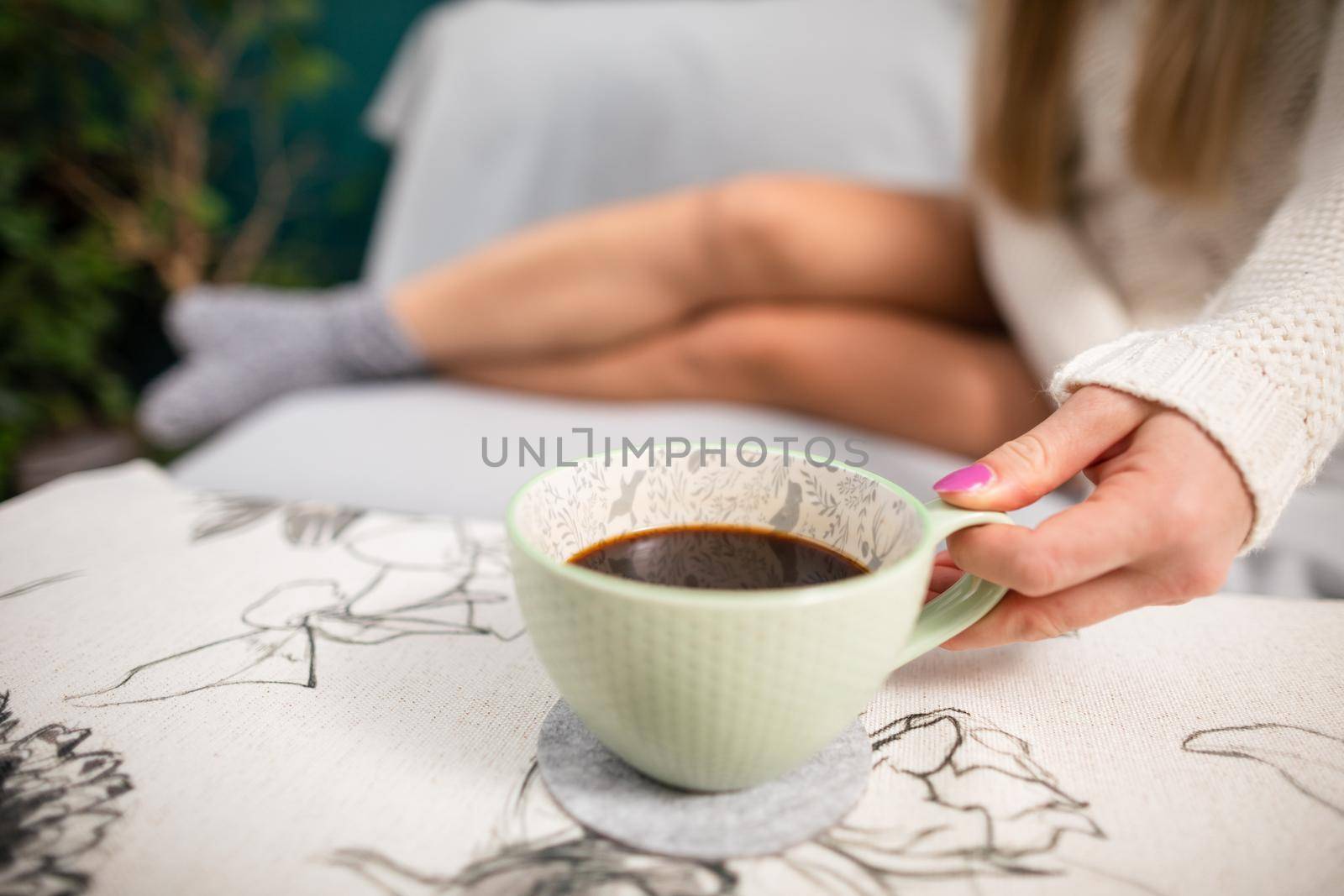 A young fetching woman with shapely legs sits on a living room sofa and in front of her holds a porcelain cup with aromatic coffee.