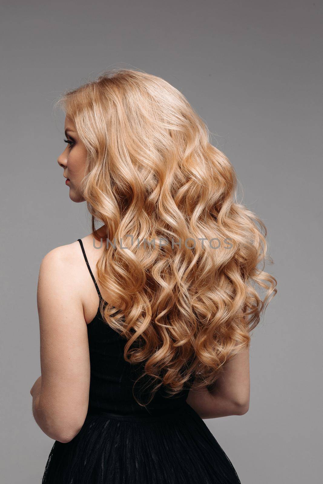 Back view of unrecognizable woman in black with volumous, blonde wavy hair over grey background. Isolated on grey. Studio. Perfect healthy hair. Hairdresser.