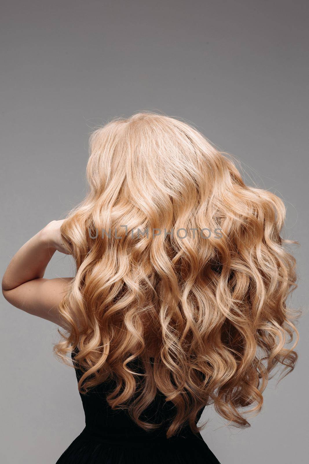 Back view of unrecognizable woman in black with volumous, blonde wavy hair over grey background. Isolated on grey. Studio. Perfect healthy hair. Hairdresser.