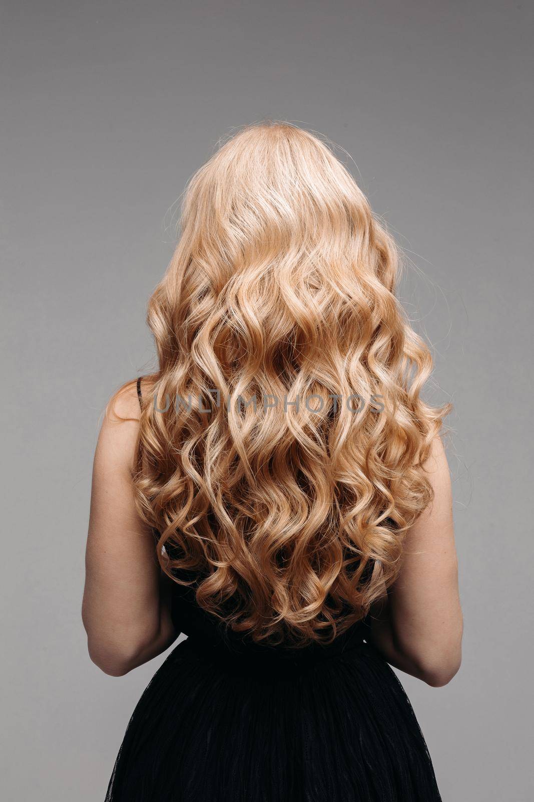 Back view of unrecognizable woman in black with volumous, blonde wavy hair over grey background. Isolated on grey. Studio. Perfect healthy hair. Hairdresser.