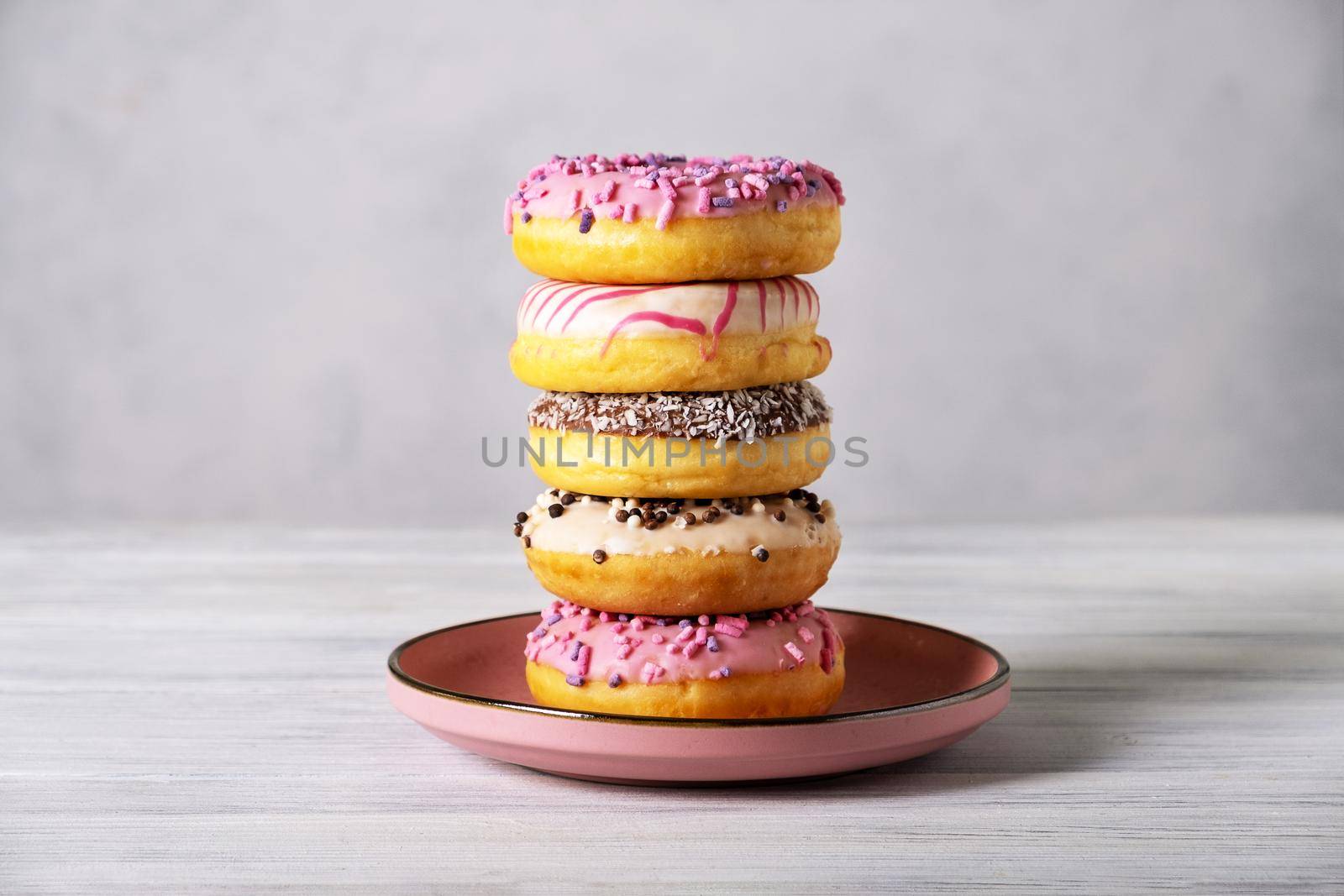 Stack of various colorful donuts on pink ceramic plate on gray background. Selective focus.