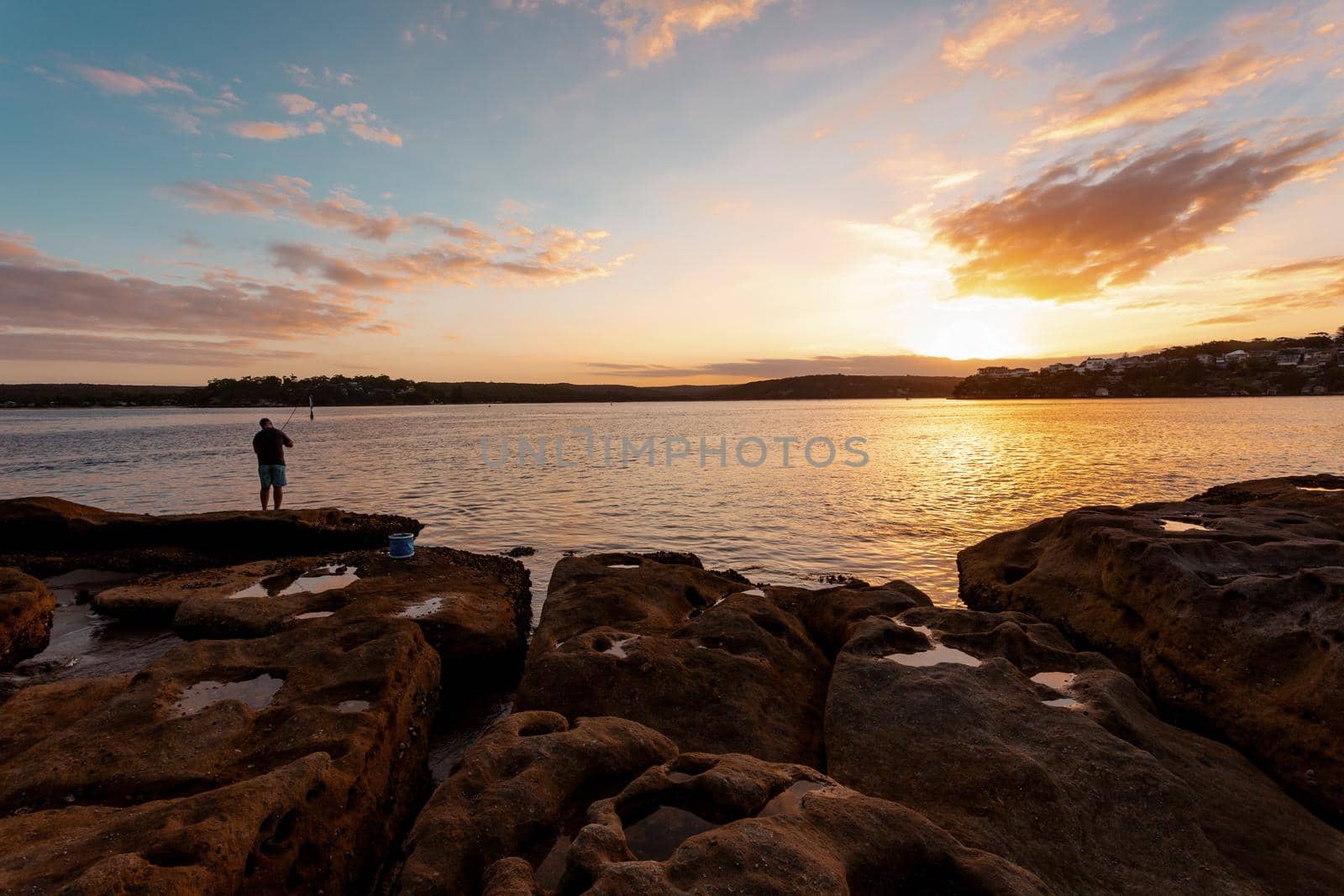 A fisherman on the rocks as the sun sets over the bay at days end.