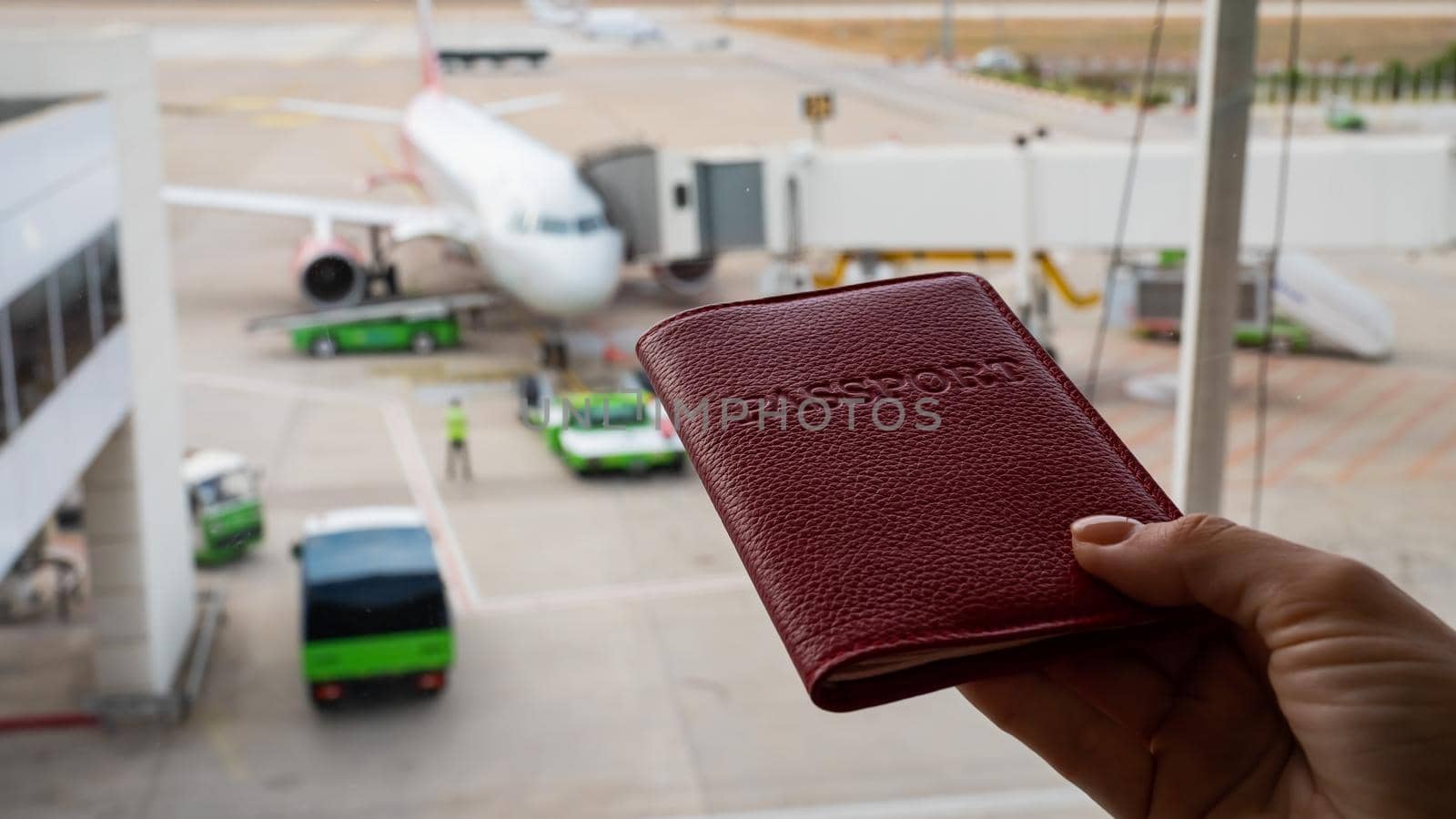 A woman holds a passport against the background of an airplane at the airport. The faceless girl is waiting for her flight and looks at the airfield through the window. by mrwed54