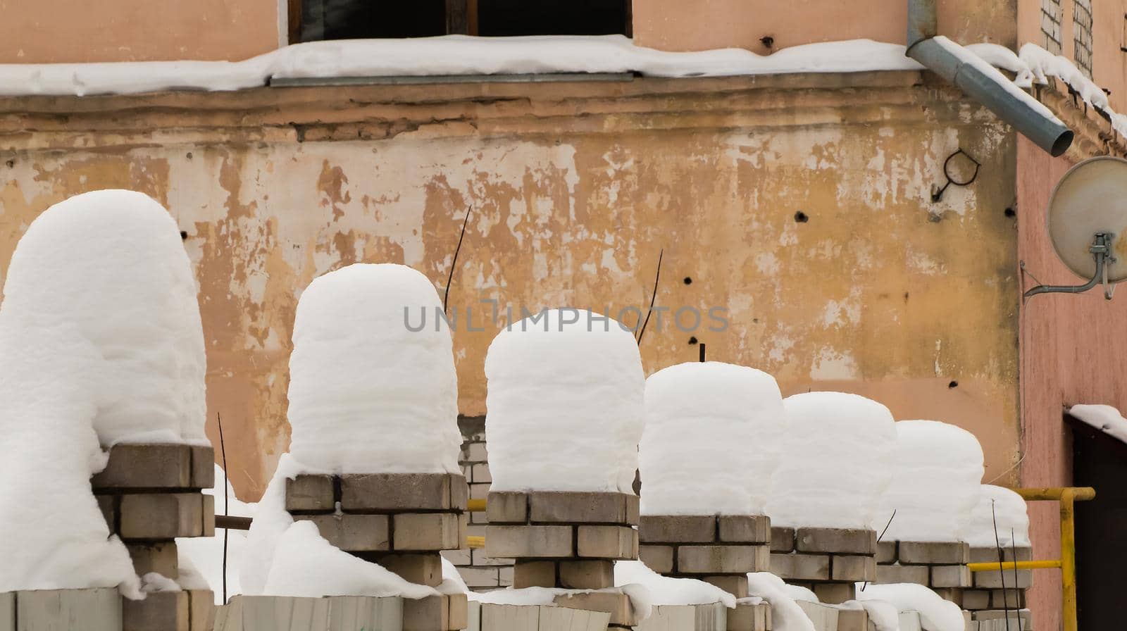 Tall beautiful columns of snow on brick pillars. An even row of small snowdrifts against the background of a shabby wall. Urban winter landscape. Cloudy winter day, soft light.