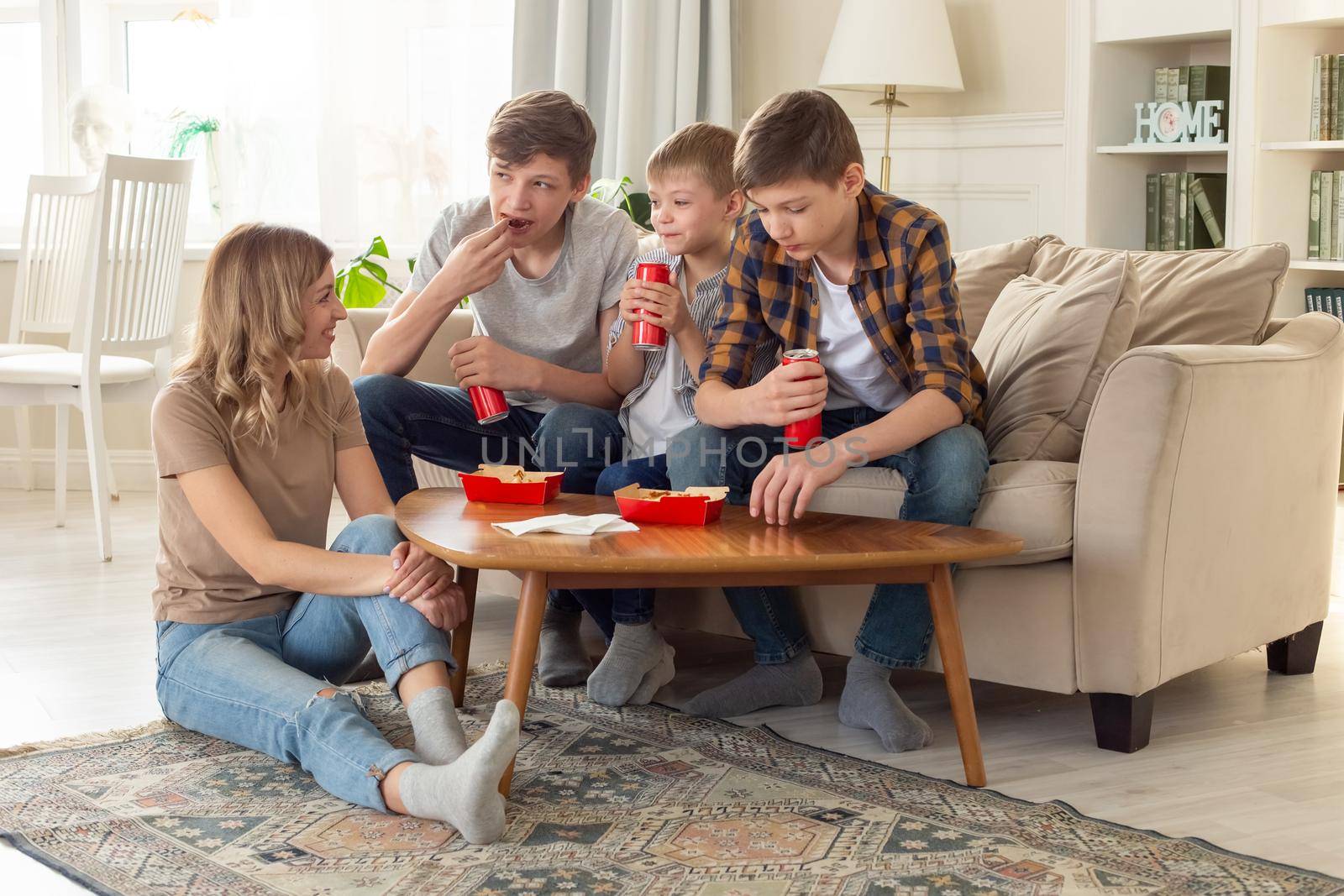 A woman and three teenage boys, are sitting on a sofa in the living room, eating fast food in red boxes and drinking drinks in red cans. copy space