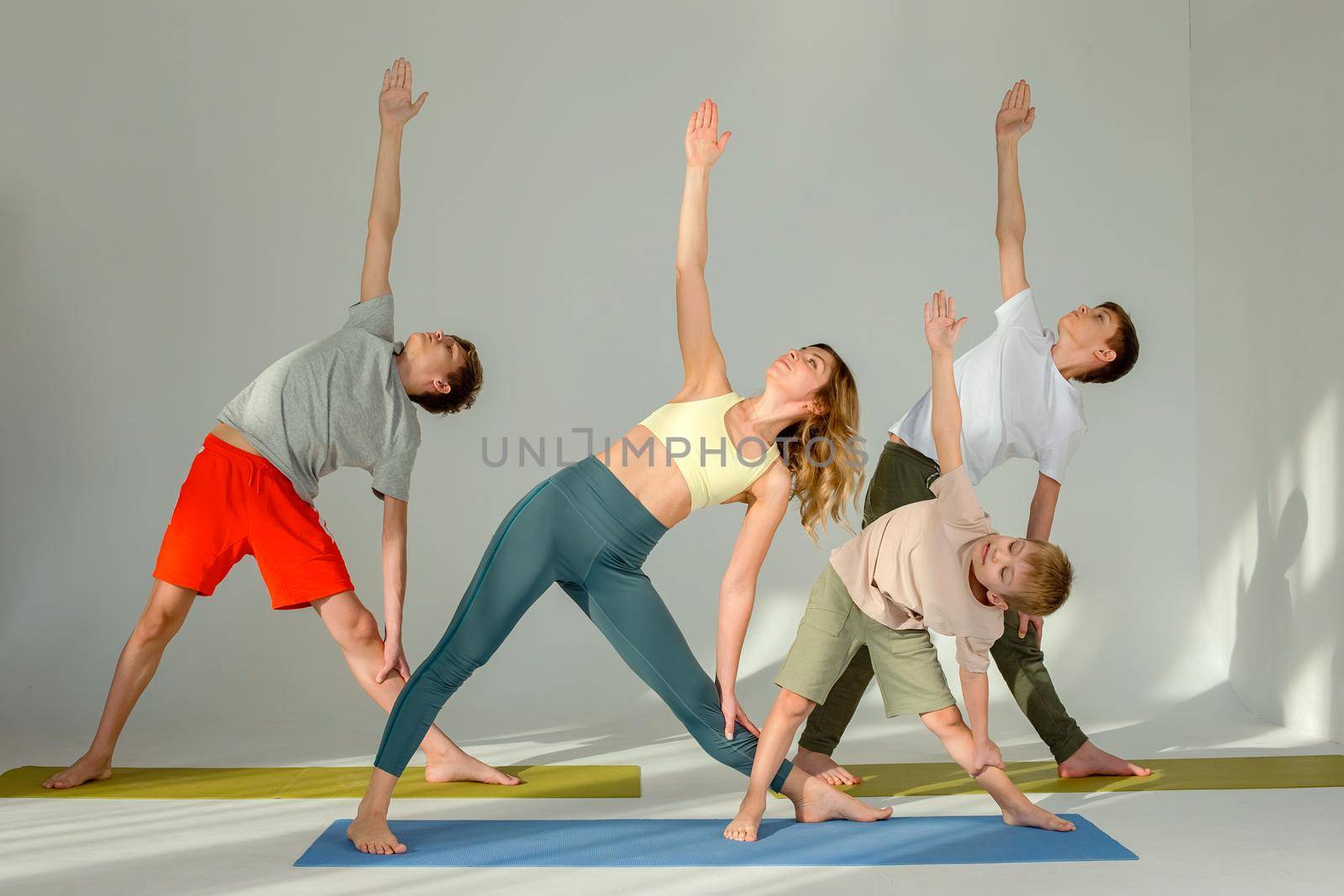 Sports family, slim woman, boy and two teenagers perform yoga exercise, stand on mat in a triangle pose, in a sunny studio.