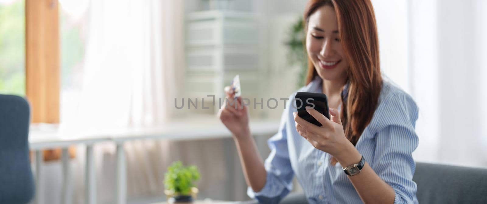 Young beautiful Asian woman using smartphone and credit card for online shopping at home with copy space. E-payment technology, shopaholic lifestyle, or mobile phone financial application concept by nateemee