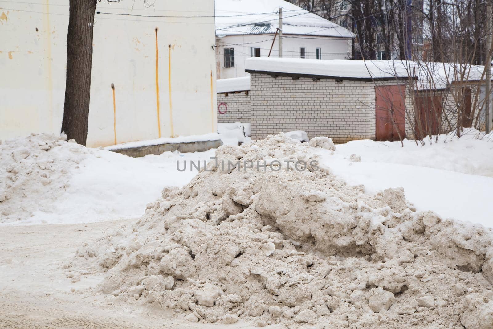 A large snowdrift by the road against the backdrop of a city street. On the road lies dirty snow in high heaps. Urban winter landscape. Cloudy winter day, soft light.