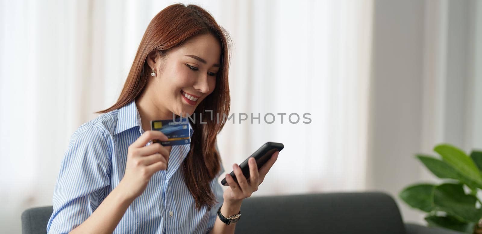 Young beautiful Asian woman using smartphone and credit card for online shopping at home with copy space. E-payment technology, shopaholic lifestyle, or mobile phone financial application concept.
