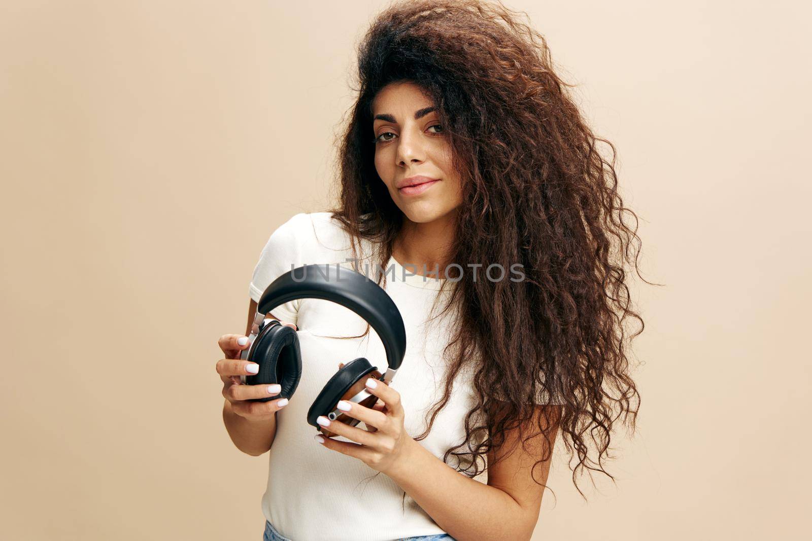 Fashionable Latin woman with afro ready to enjoying cool songs, hold wireless headphones, close eyes, stay isolated near beige wall. Copy free space for design ad. Fashion IT Seasonal Sale concept by SHOTPRIME