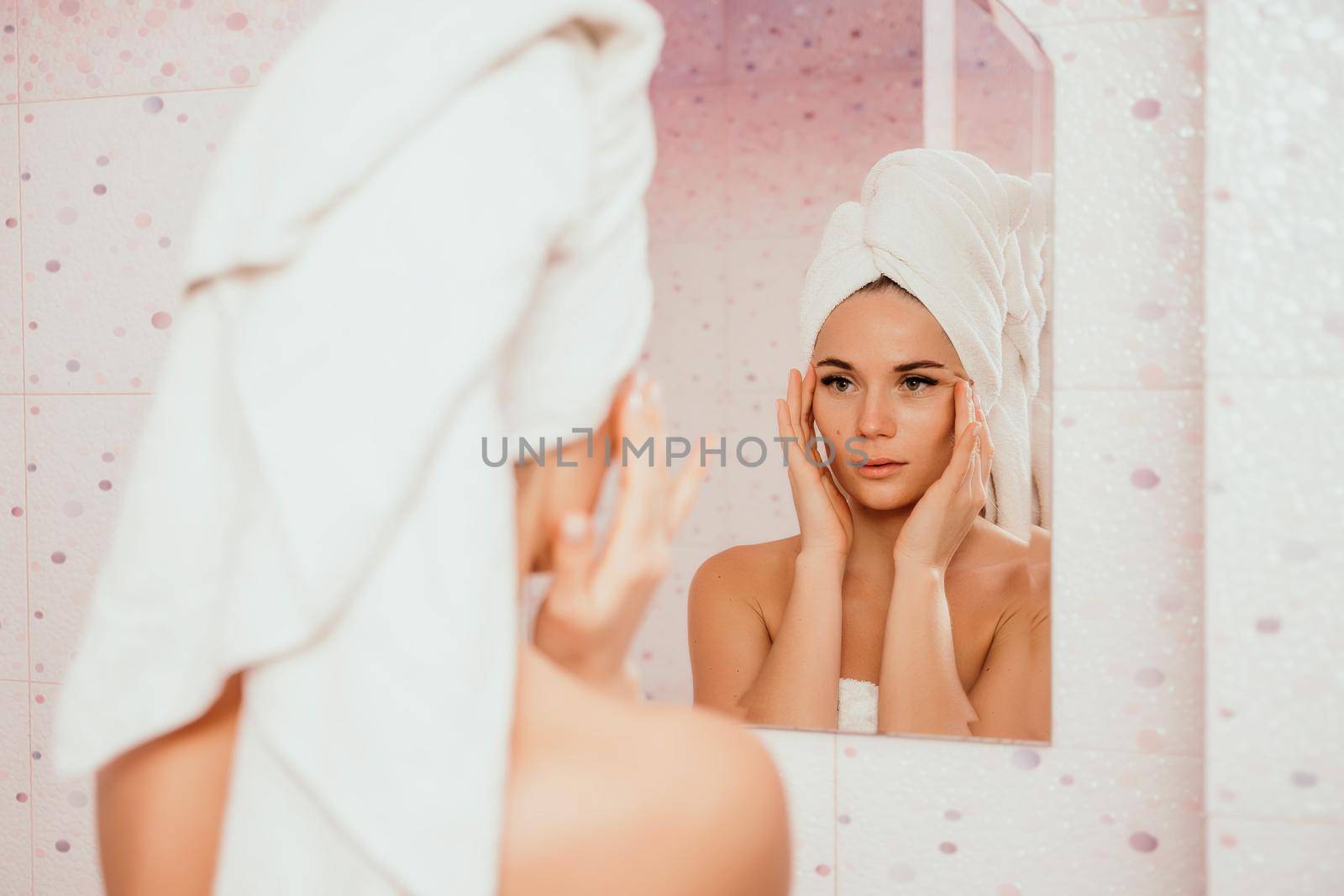 Young beautiful woman using face cream moisturizing lotion after bath. Pretty attractive girl in a towel on her head stands in front of a mirror in a home bathroom. Daily hygiene and skin care.