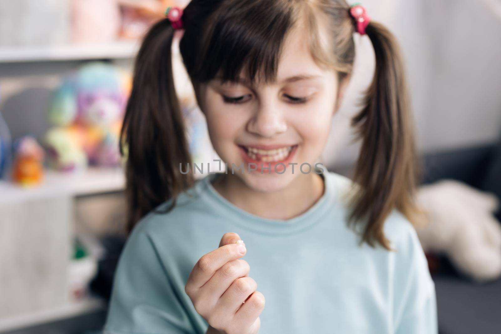 Portrait of a baby girl with a toothless smile. Shows a ripped tooth into the camera in an extended hand. Shift focus from tooth to face. Dental medicine or temporary teeth health care concept.