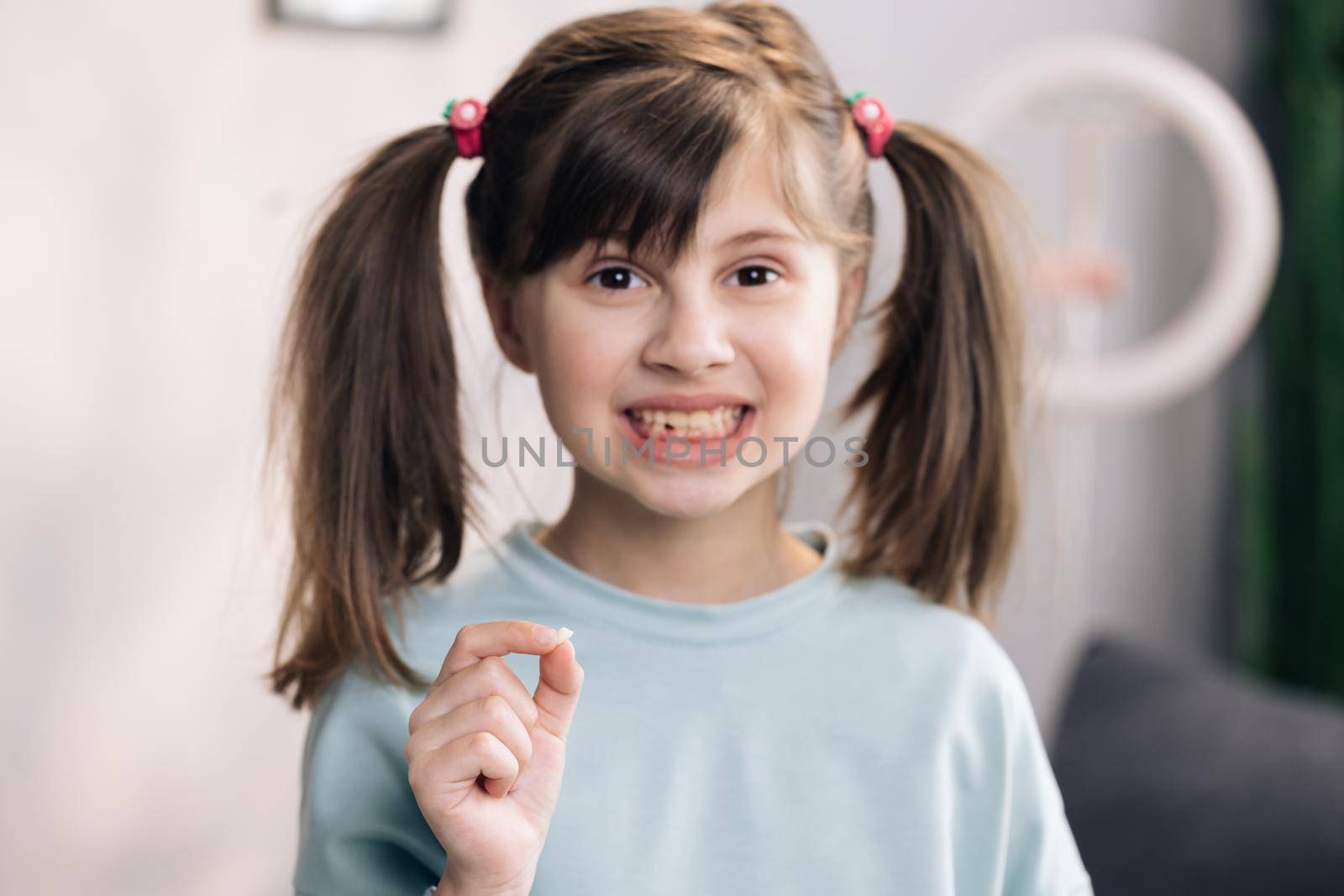 Happy girl holding her fallen tooth in hand. Portrait of cute little child girl is rejoices and showing her lost milk tooth and smiling to camera of toothless mouth while she standing in bedroom.
