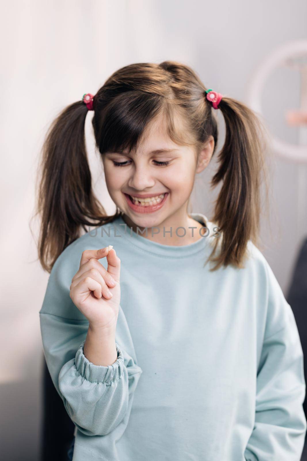 Cute little child girl is rejoices and showing her lost milk tooth and smiling to camera of toothless mouth while she standing in bedroom. Happy girl holding her fallen tooth in hand. by uflypro