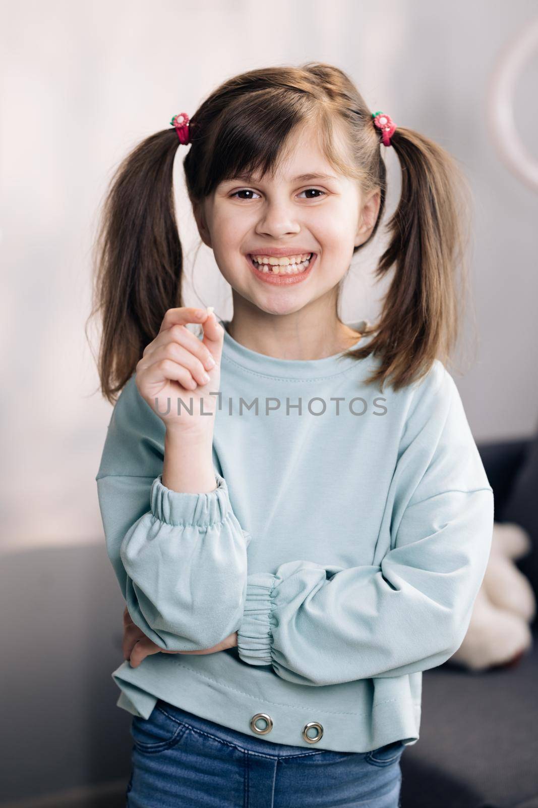Portrait of cute little child girl is rejoices and showing her lost milk tooth and smiling to camera of toothless mouth while she standing in bedroom. Happy girl holding her fallen tooth in hand. by uflypro