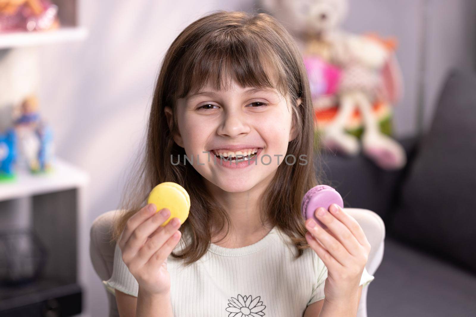 Happy smiling face of a little girl covering her eyes with macaroons on a colored home background. Being happy. Teen girl plays with dessert macarons, holding the cookies like glasses around the eyes.