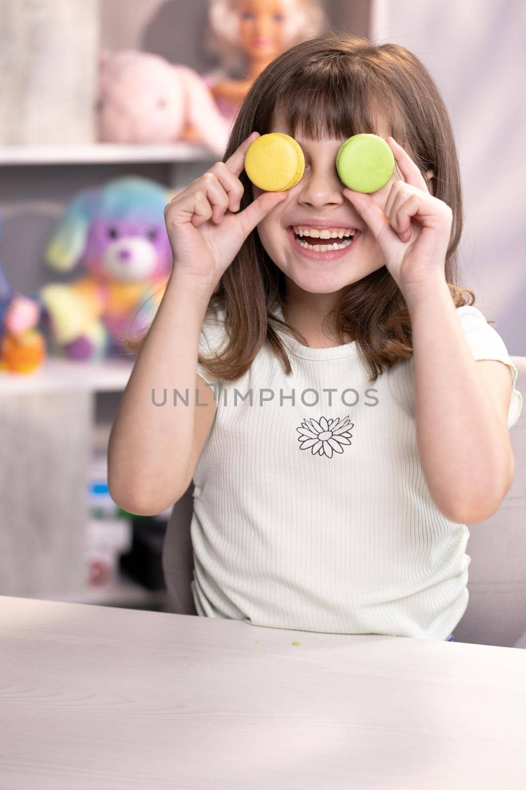 Happy smiling face of little girl covering her eyes with macaroons. Teen girl plays with dessert macarons, holding the cookies like glasses around the eyes and throws his hands to the sides.