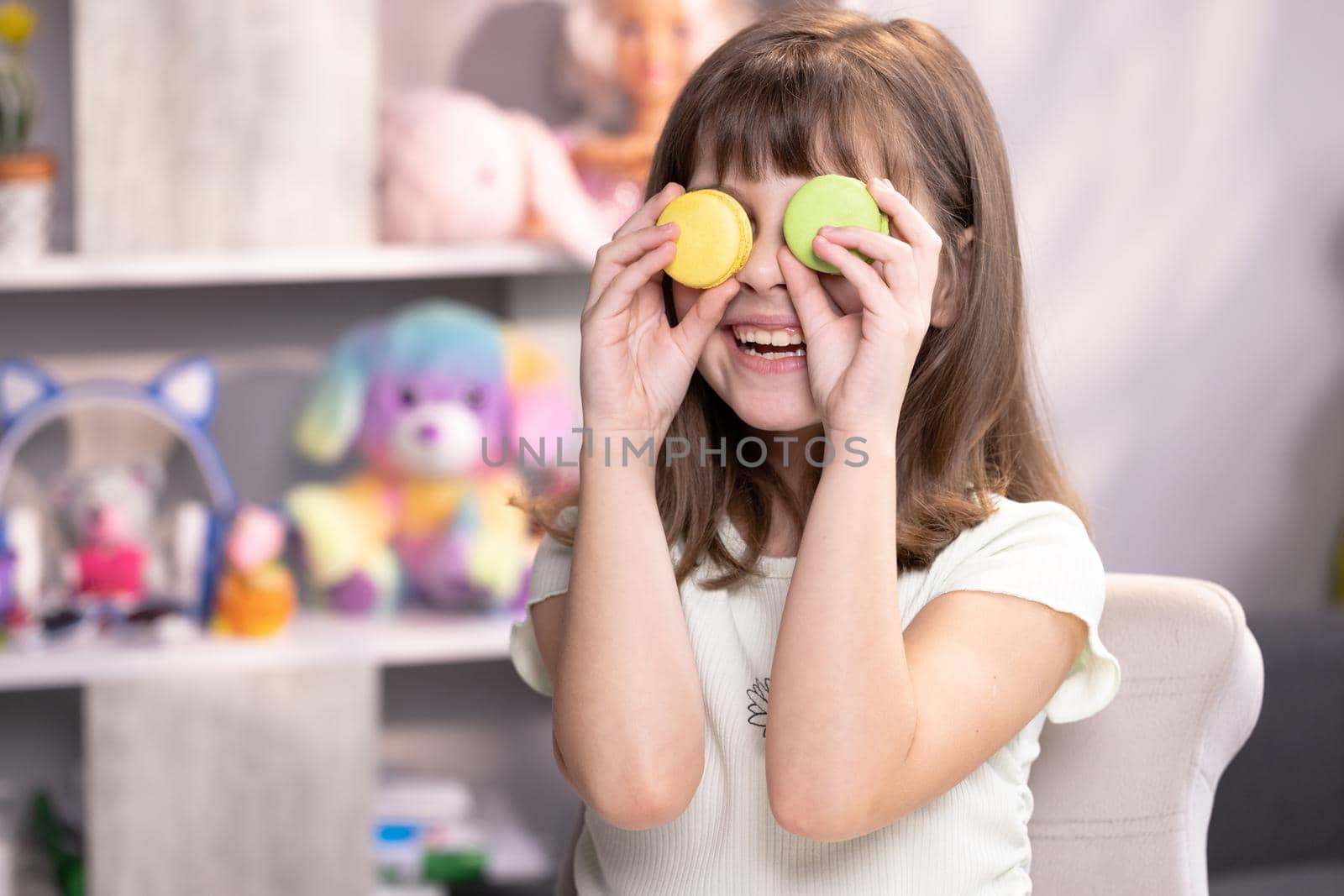 Happy smiling face of little girl covering her eyes with macaroons. School girl plays with dessert macarons, holding the cookies like glasses around the eyes and throws his hands to the sides.
