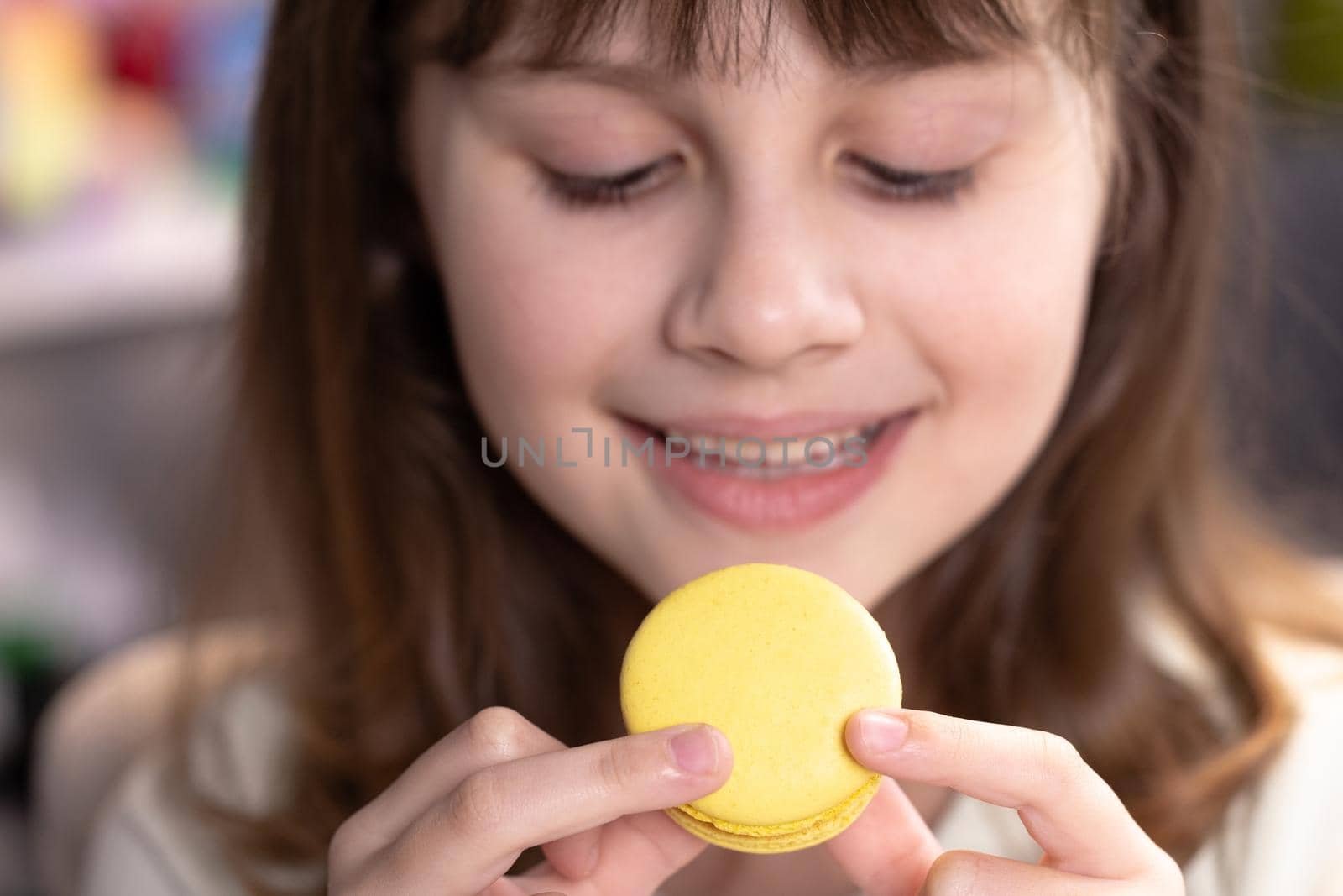 Attractive brunette school girl on a home background, licks her lips, eating one yellow macaron, closes her eyes with pleasure. Dessert person, sweet tooth, gourmet. Feeling good, delicious by uflypro