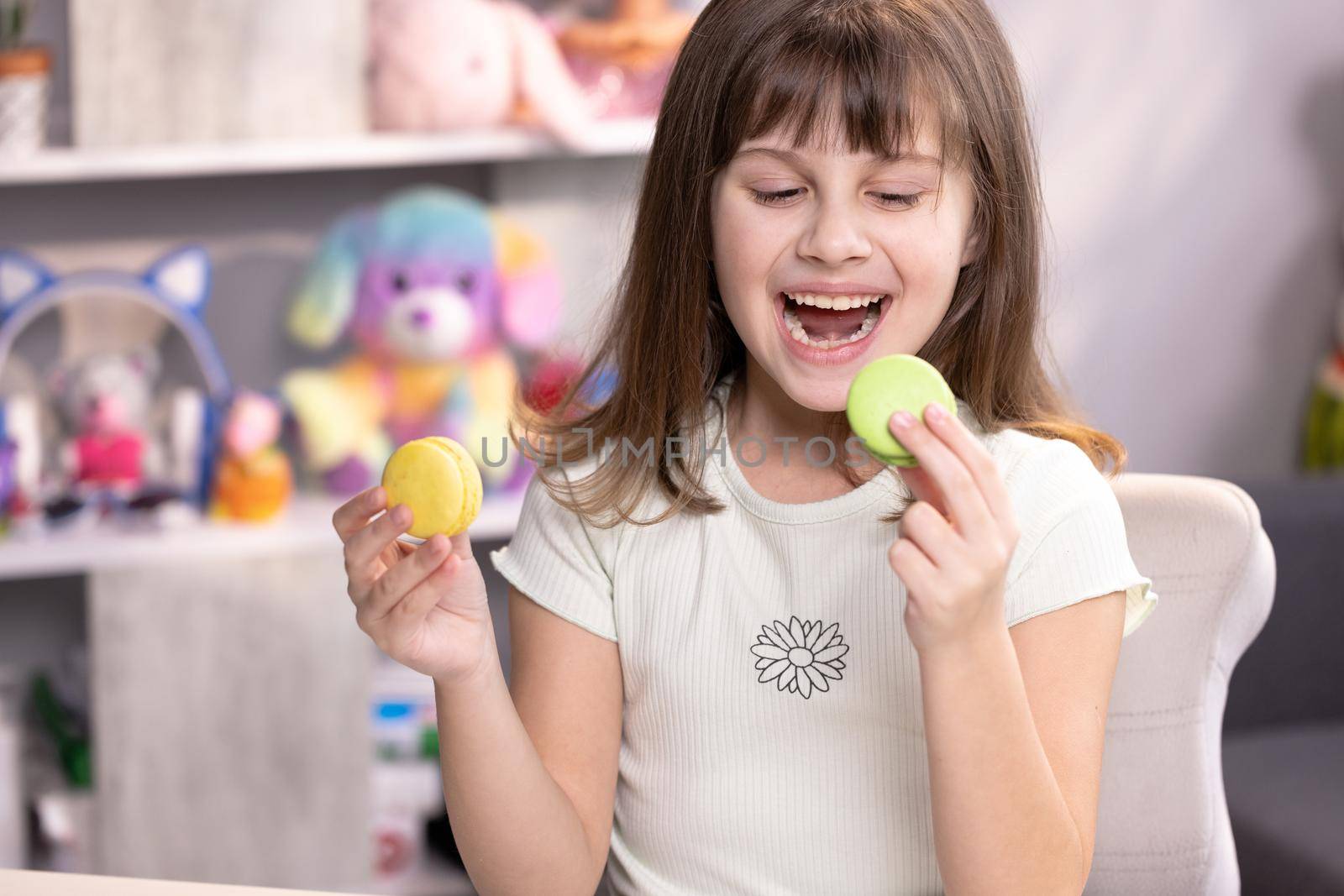 Teen girl plays with dessert macarons, holding the cookies like glasses around the eyes. Happy smiling face of a little girl covering her eyes with macaroons on a colored home background. Being happy by uflypro
