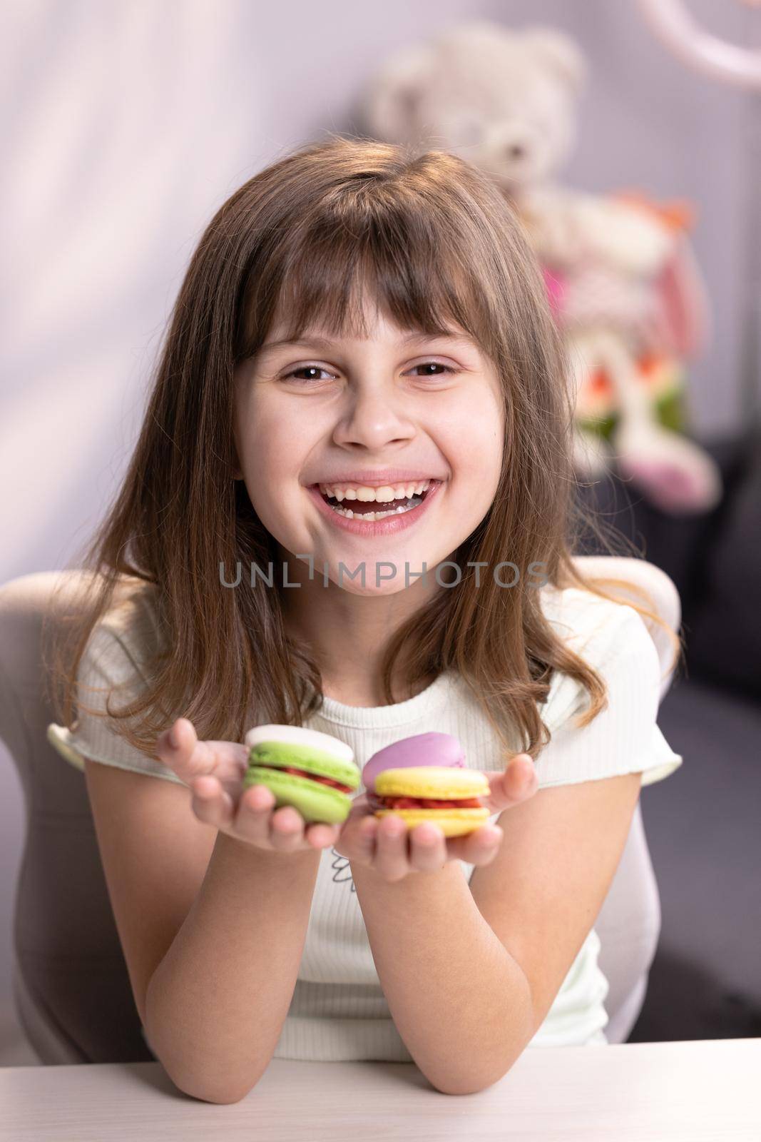 Attractive smiling cute school kid girl holds four tasty cookies in her hands and look at camera. Traditional French multicolored macaroon. Food concept by uflypro