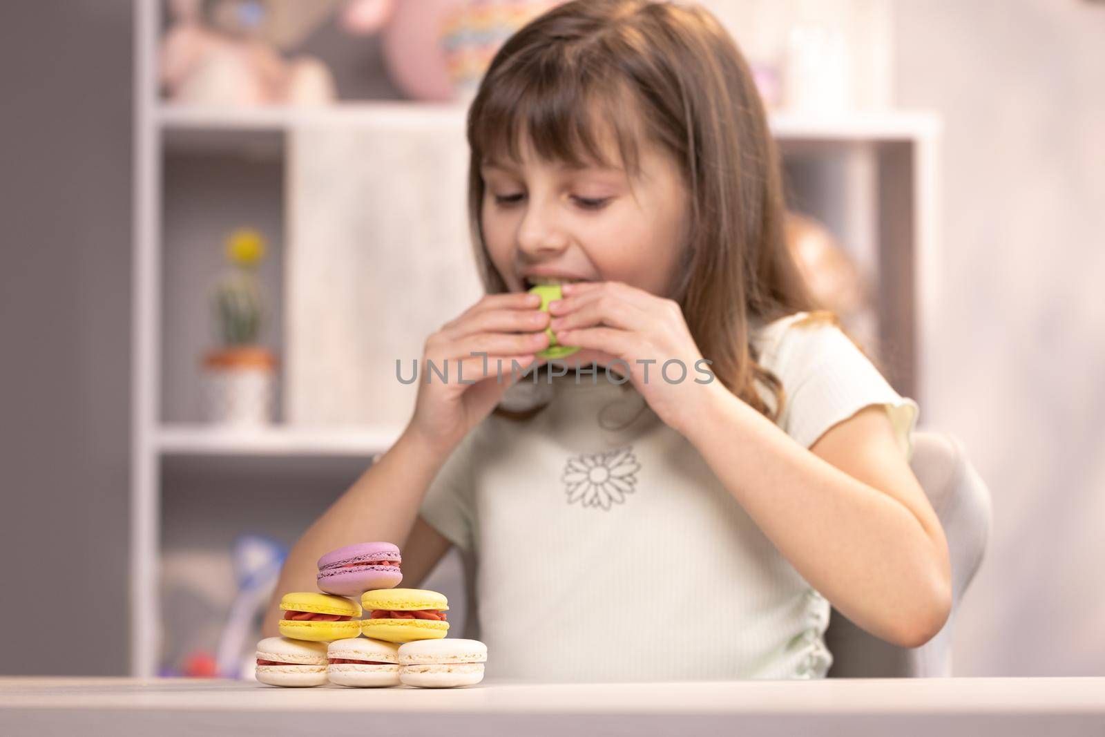 Happy smiling face of a little girl plays with dessert macarons on home background. Brunette kid girl bites pistachio green macaron cookie. Dessert person, sweet tooth, gourmet.