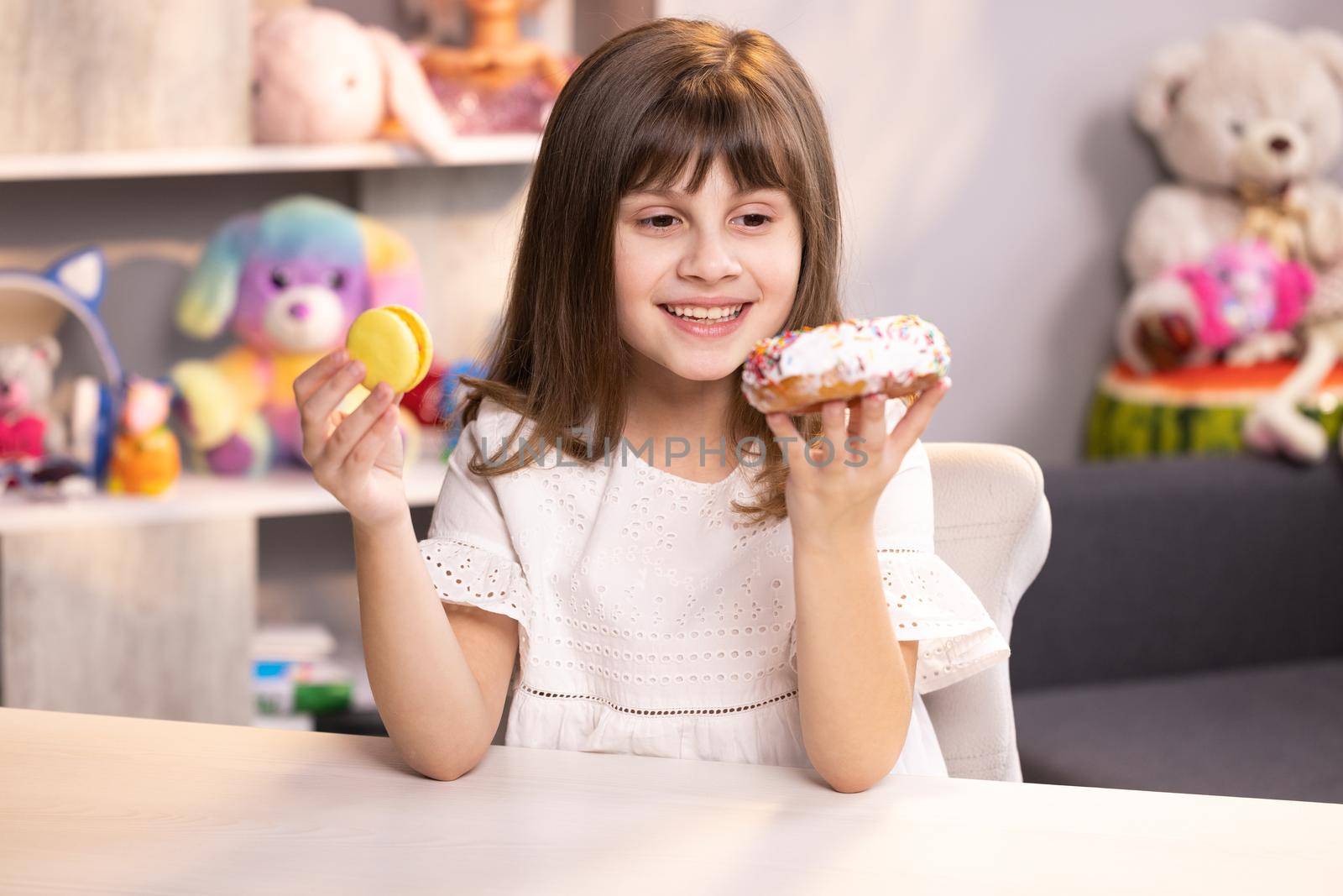 Lovely joyful school girl can not choose between a white donut and yellow fresh macaroon. Funny teenager girl having fun with donut and macaron at modern home. Sweet tooth. Unhealthy food concept.